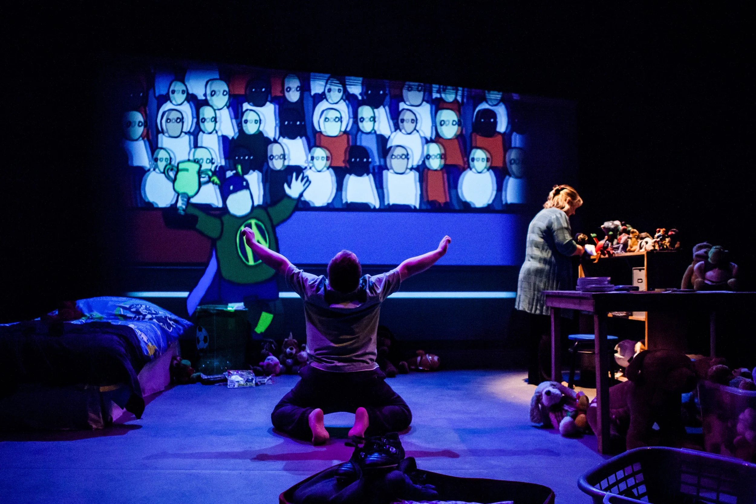 Actor Nathan Bessell is kneeling on the floor with his arms outstretched towards an animation of a crowd of people and a superhero. The colours are purple, blue, red and green.