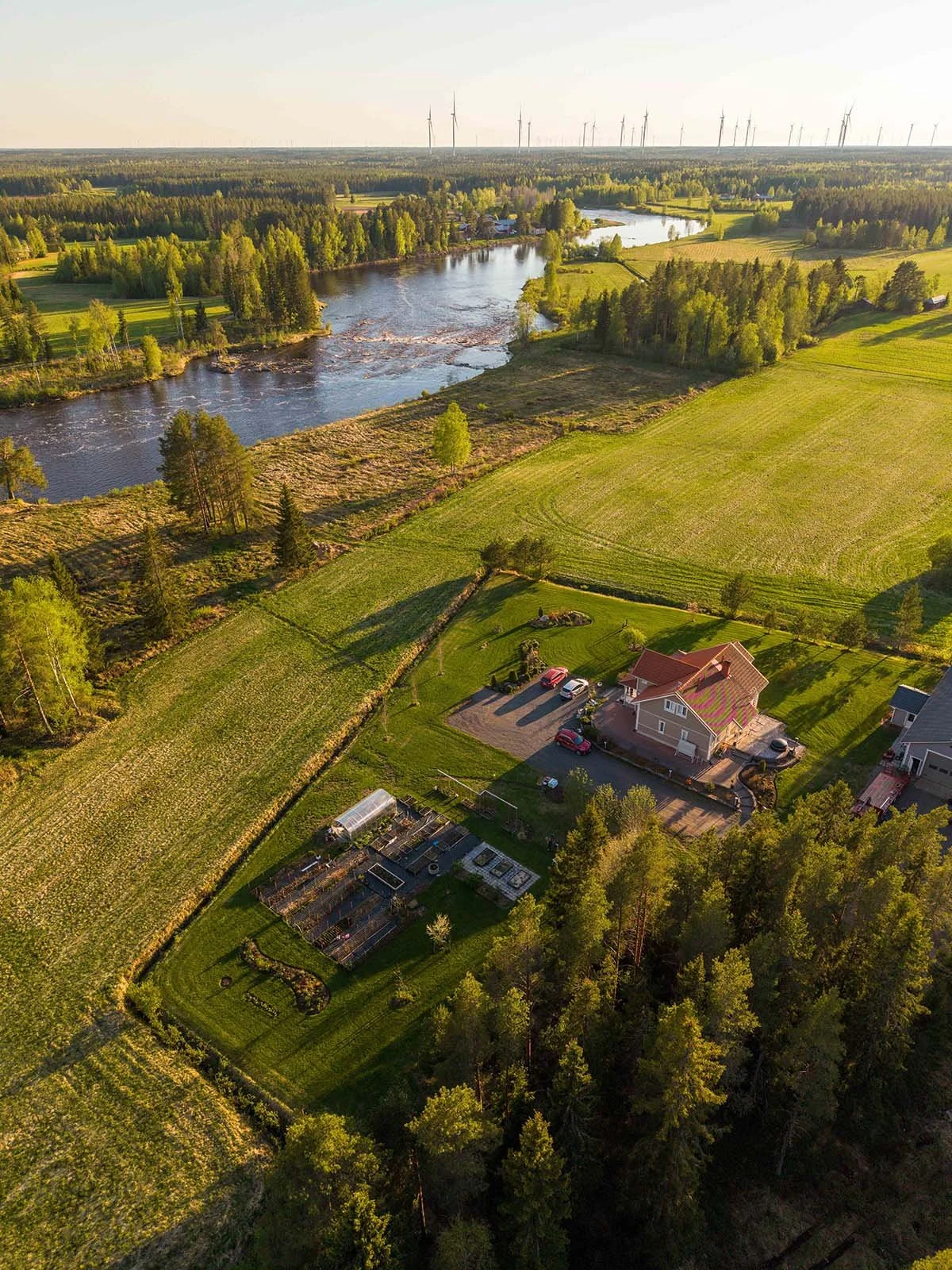 Dronekuva, joka näyttää vehreän maalaismaiseman, joen ja talon päiväsaikaan.