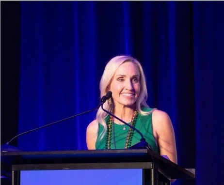 A woman with long blonde hair smiling while speaking at a podium on stage with a blue curtain backdrop.