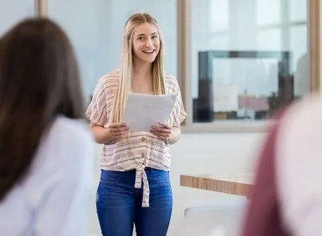 A young woman smiling and holding papers, standing in a bright office or classroom, with blurred people in the foreground.