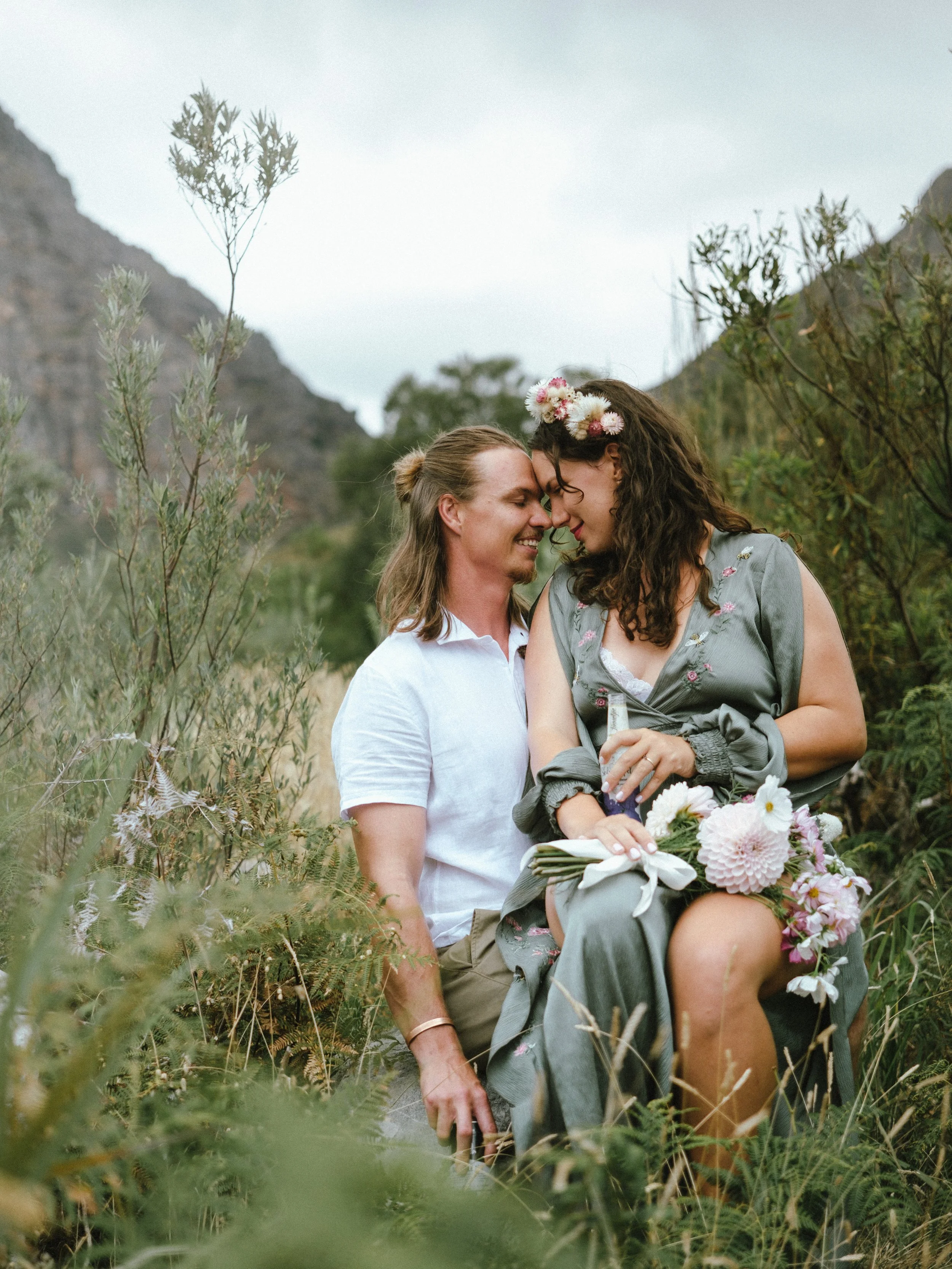 A couple sitting close together in a grassy, mountainous outdoor setting, with their foreheads touching and eyes closed. The woman is wearing a gray dress with floral embroidery and a flower crown, holding a bouquet of flowers. The man is dressed in a white shirt and beige pants, smiling gently.