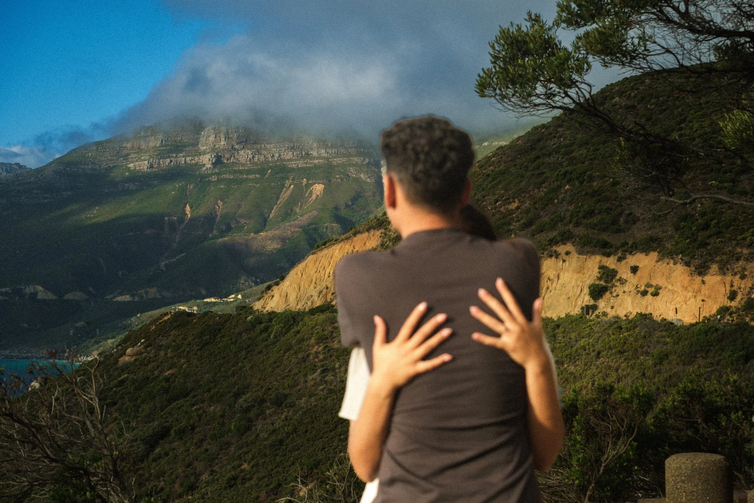 A couple embraces each other with an ocean and green hillside in the background, during daytime.