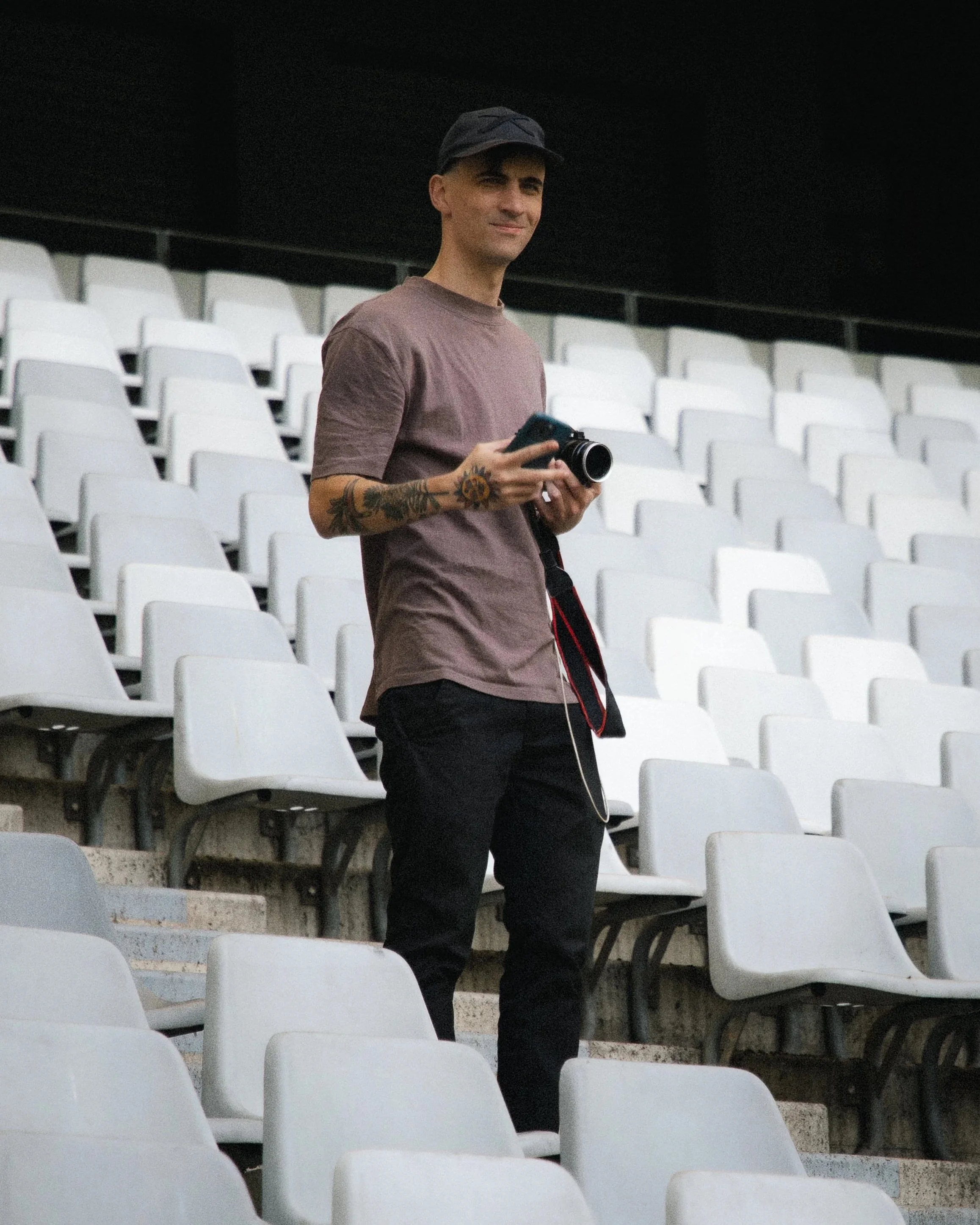 A young man standing in an empty stadium, holding a camera, wearing a dark cap, a brown t-shirt, and black pants.