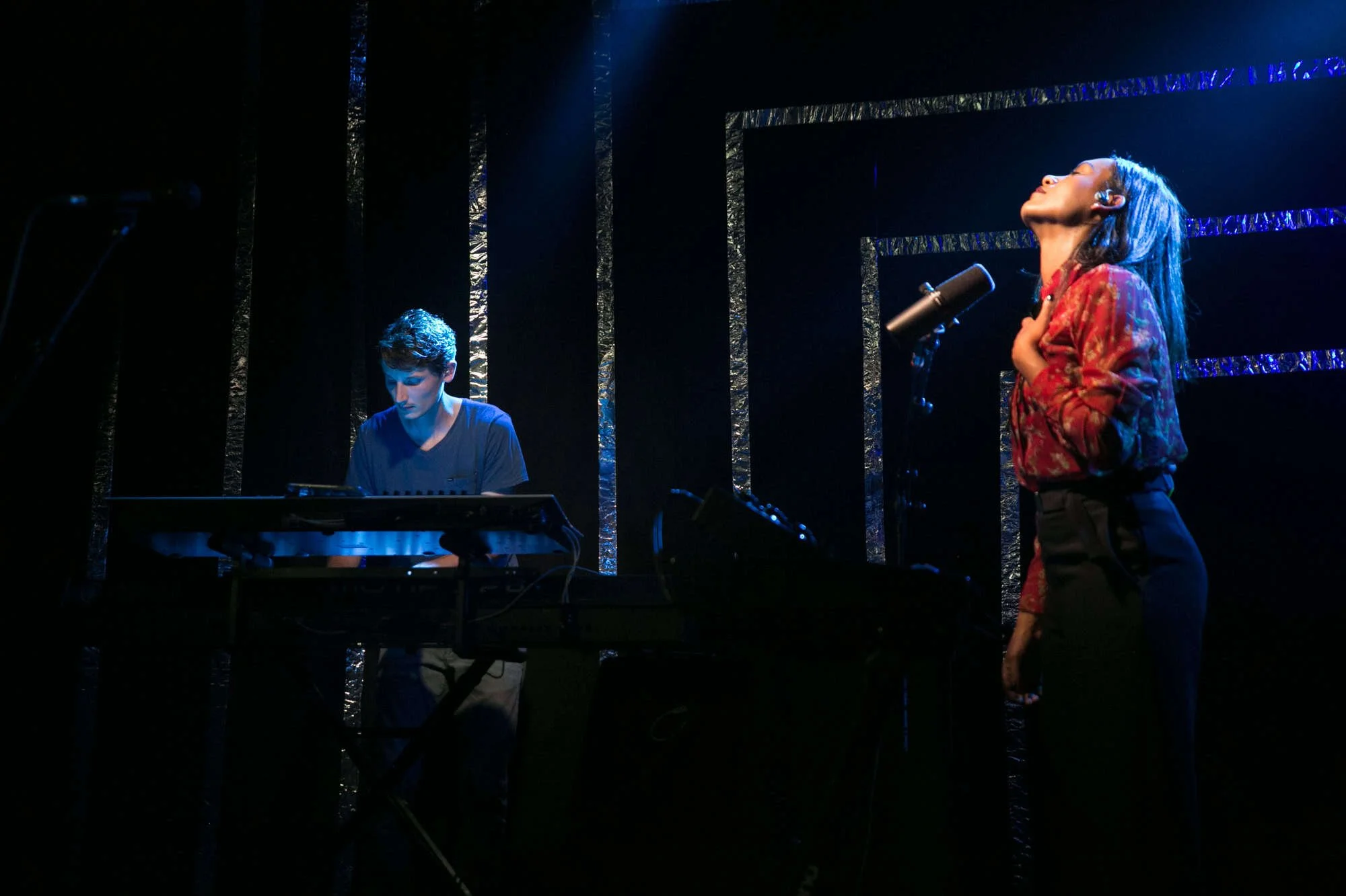 Chanteuse et claviériste en plein concert à Genève devant une installation scénographique ingénieuse. L'arrière-plan est structuré par des ruban adhésifs argentés suspendus qui accrochent la lumière, détournement de matériaux simples