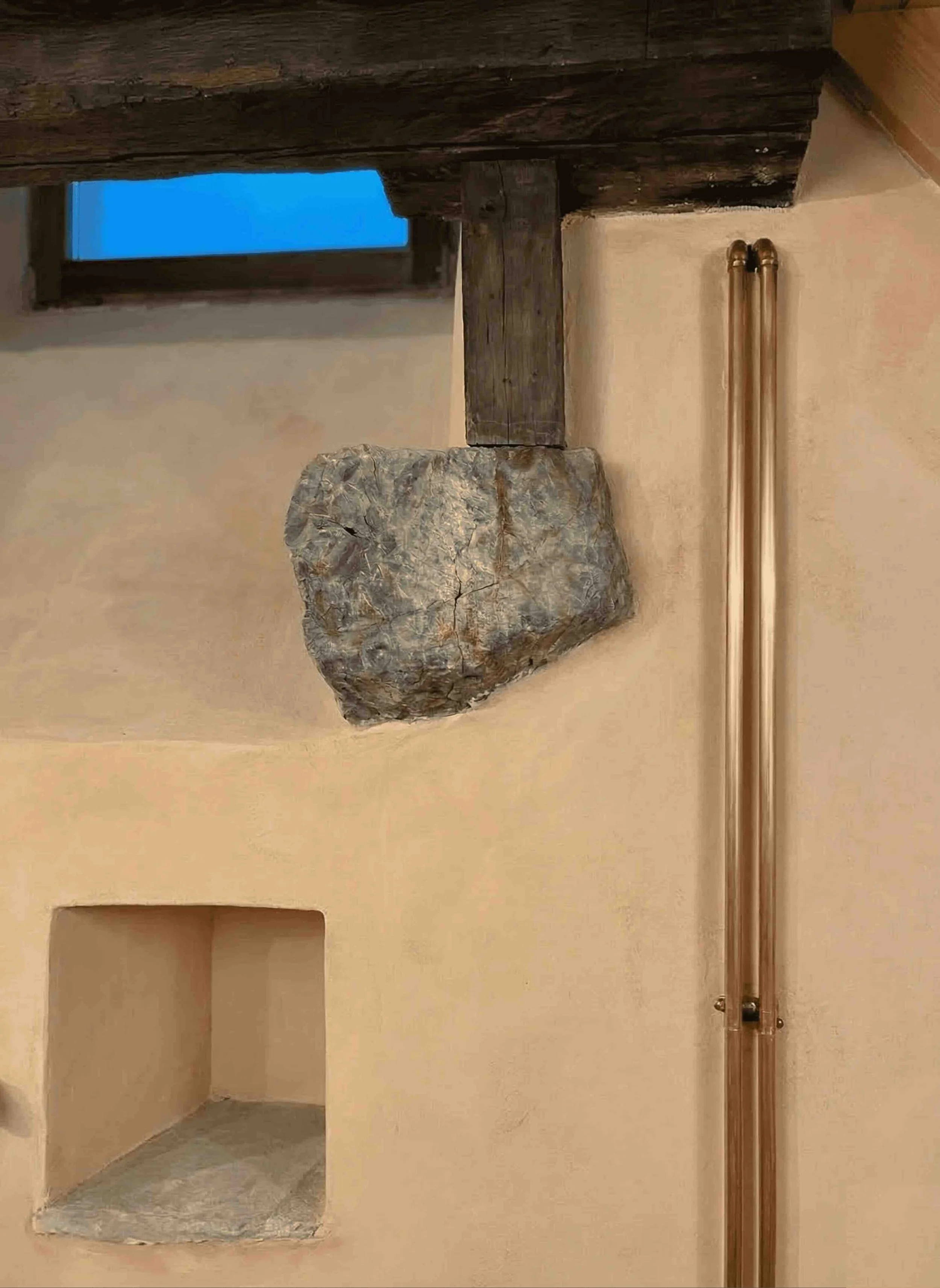 Détail architectural d'une rénovation à Genève : mise en valeur d'une pierre brute d'origine sous poutre et d'une ancienne tuyauterie en cuivre verni sur un mur à la chaux.