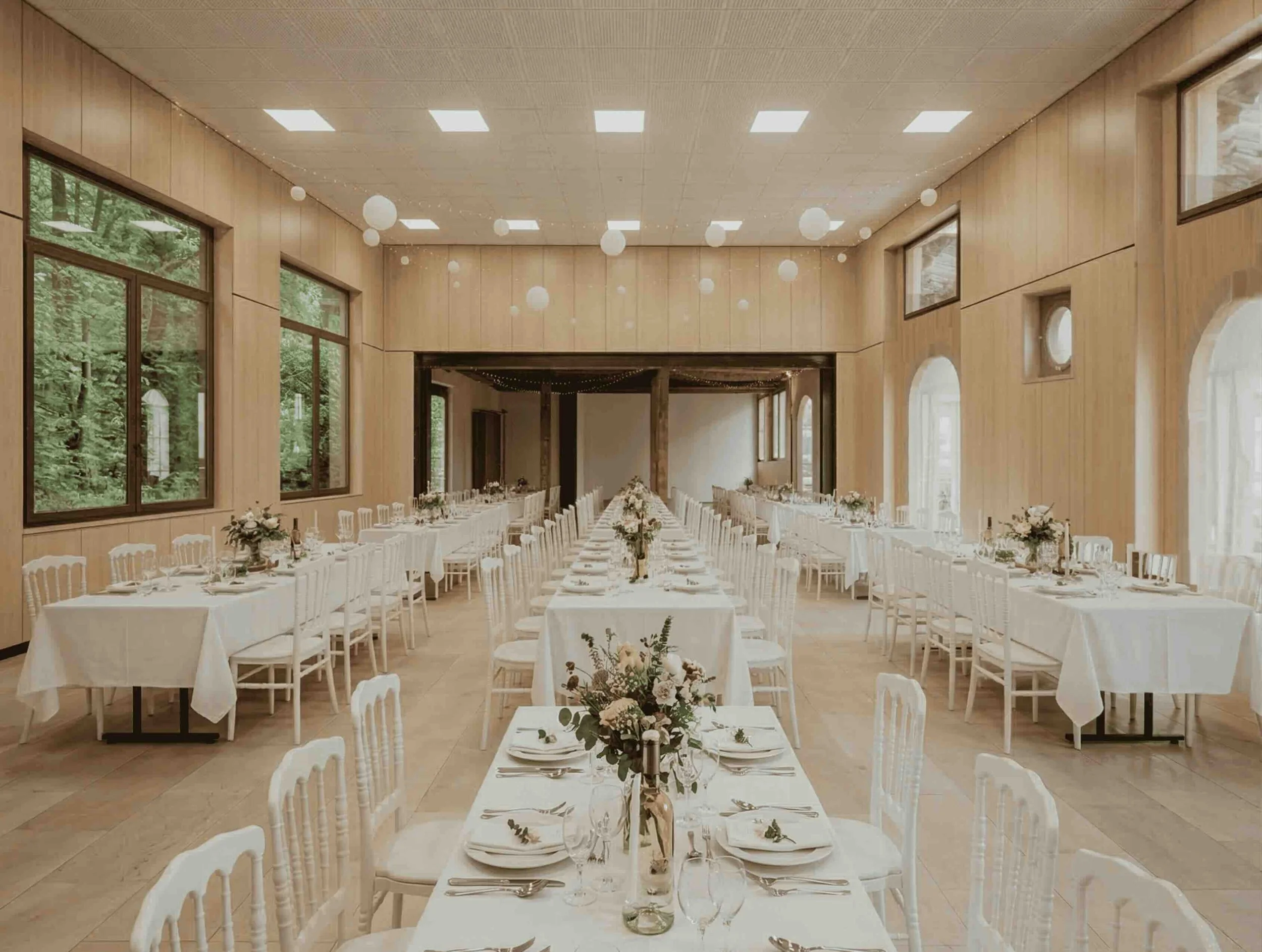 Vaste salle de mariage lumineuse habillée de boiseries claires à Genève. De longues tables de banquet occupent ce volume monumental et unifié, rendu possible par la suppression d'un mur porteur central lors de la transformation des anciens garages.
