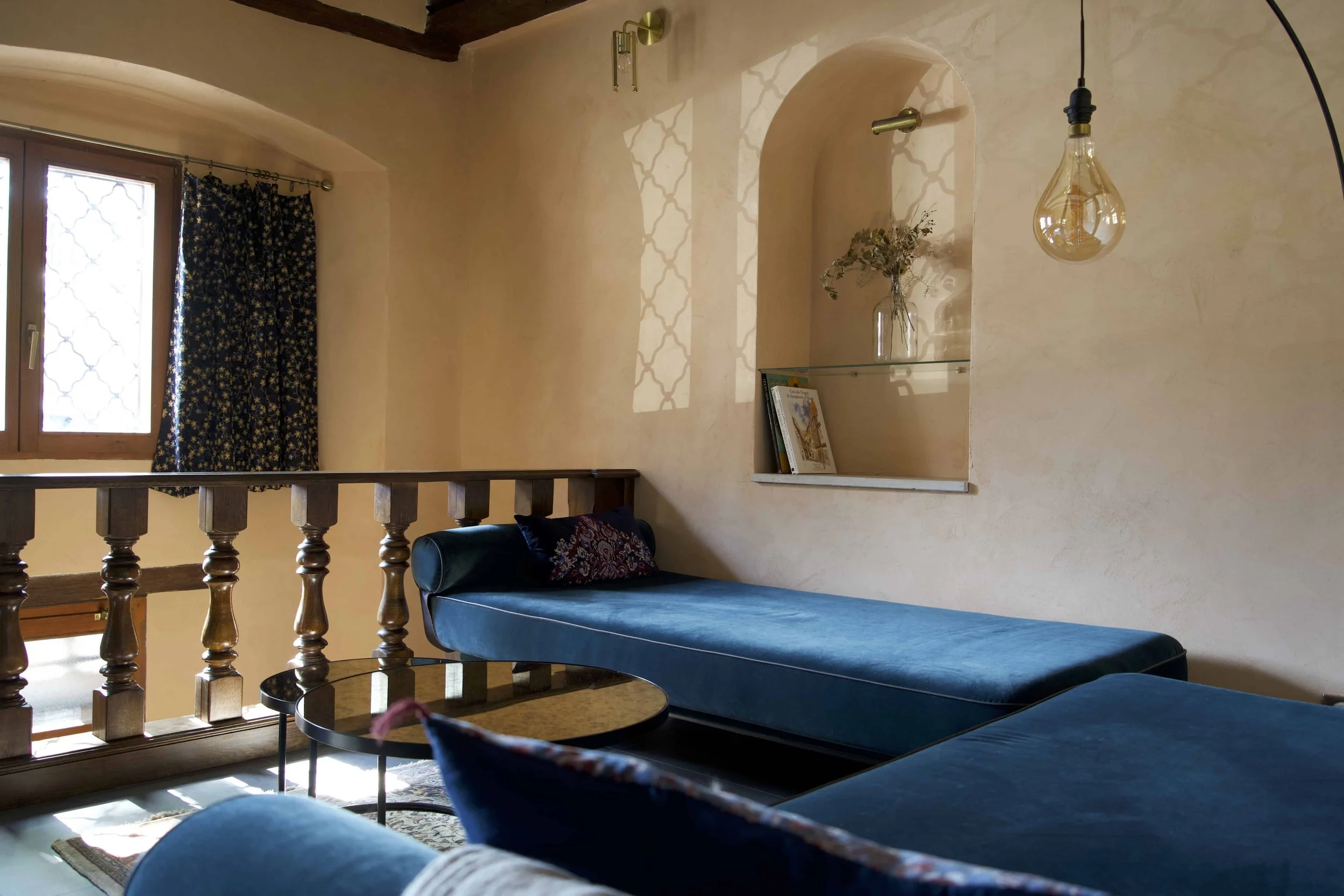 Design d'intérieur sur-mesure par un architecte à Genève : aménagement d'un salon sous poutres apparentes, où le mobilier audacieux en velours bleu profond contraste avec les tons minéraux des murs enduits.