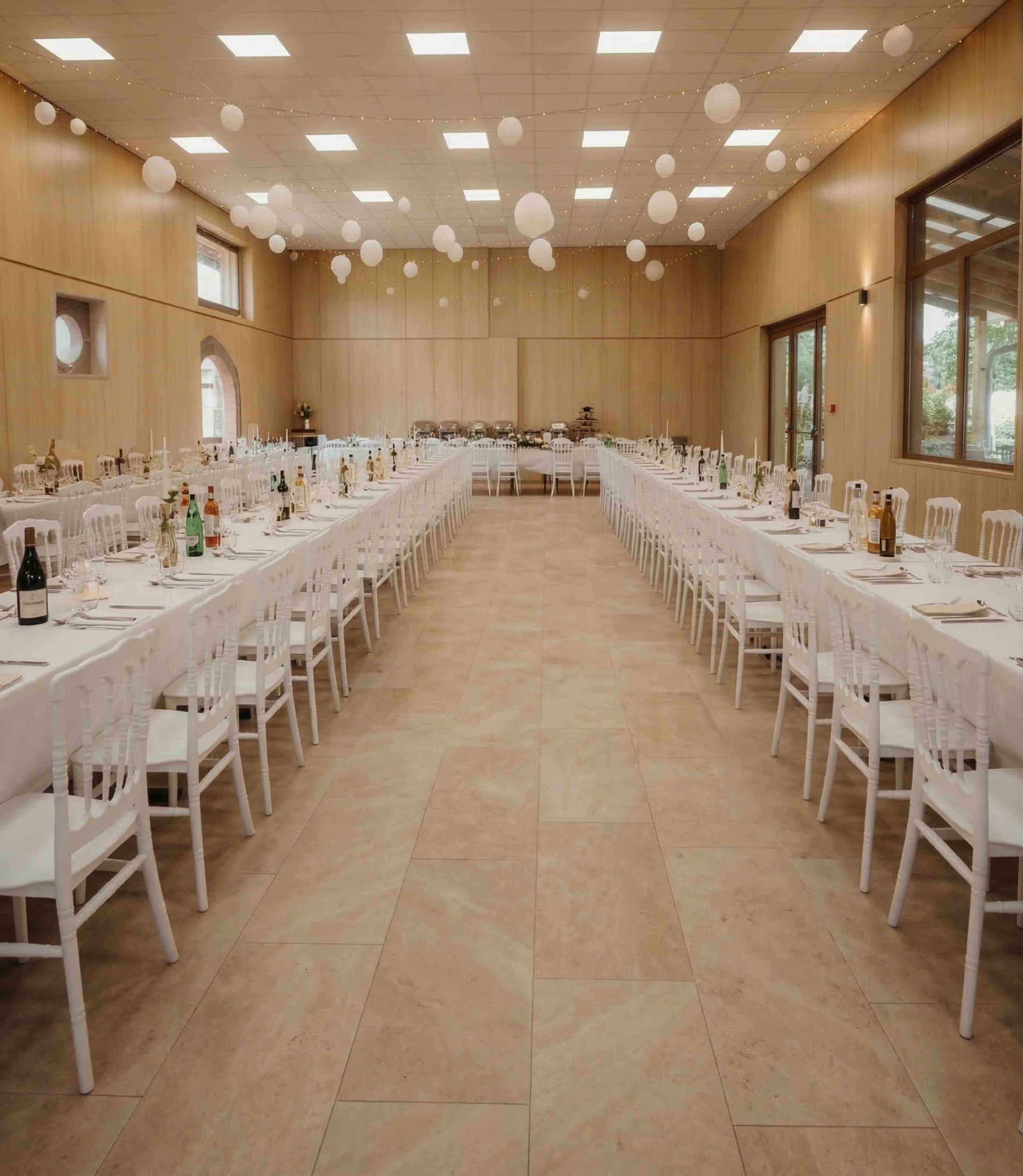 Intérieur élégant et lumineux d'une grande salle de réception pour mariages et événements à Genève. L'image montre de longs banquets dressés avec des chaises Chiavari blanches sur un sol en pierre avec mur en bois clair. Transformation d'un hangar