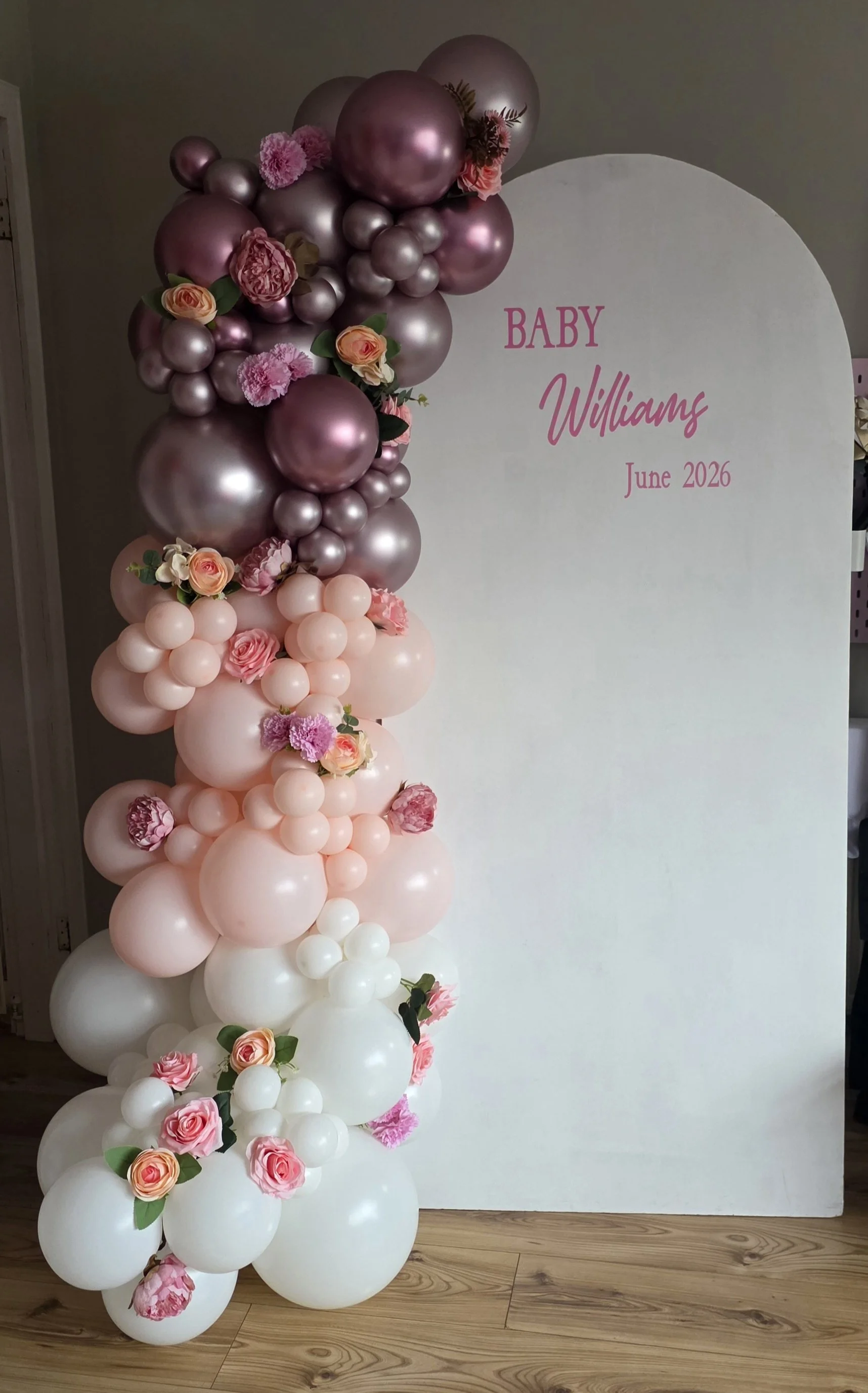 Decorative balloon arch in shades of pink, purple, and white with floral accents, standing next to a white backdrop that reads 'Baby Williams June 2026' in pink text.