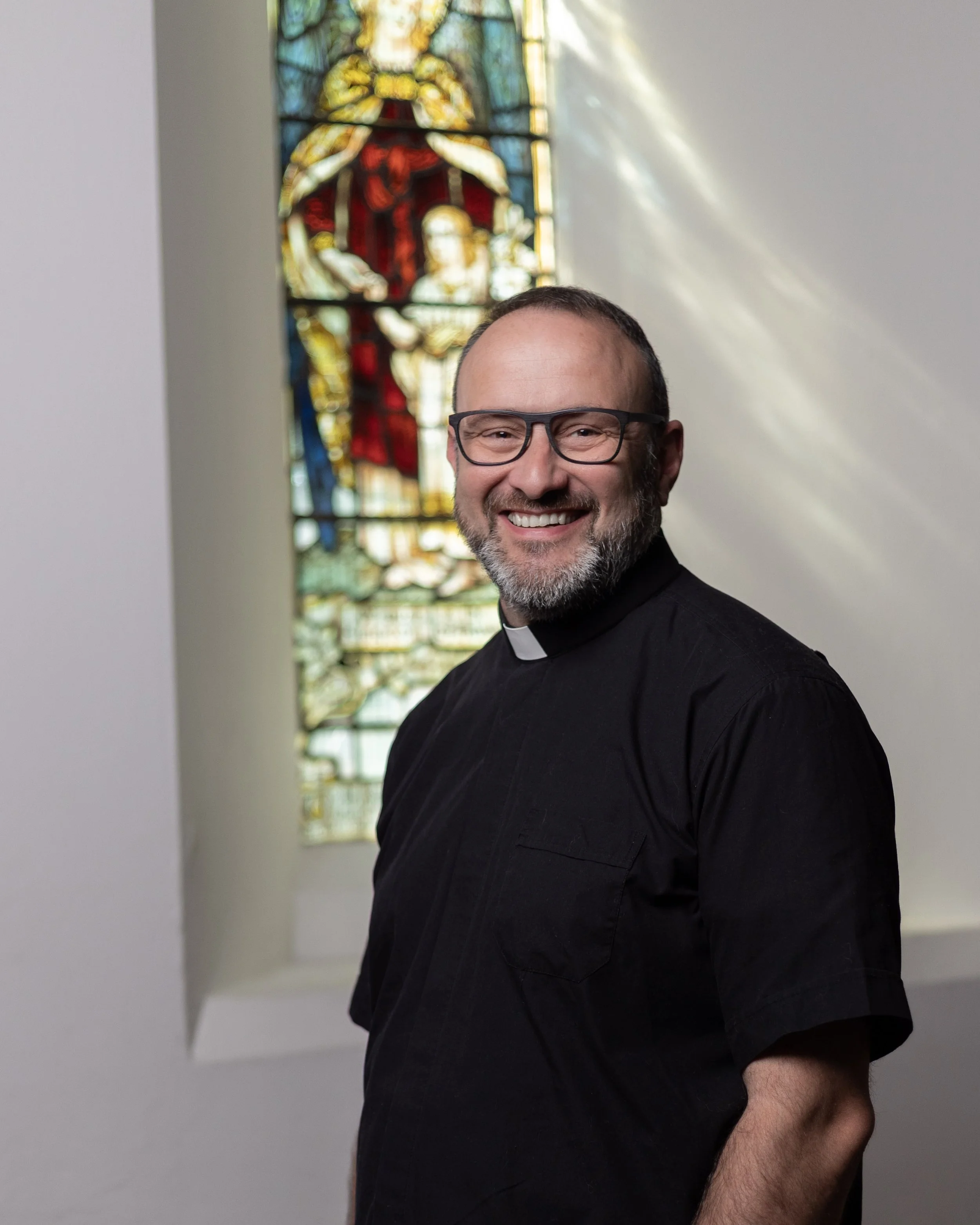A smiling man dressed as a priest standing in front of a colorful stained glass window.