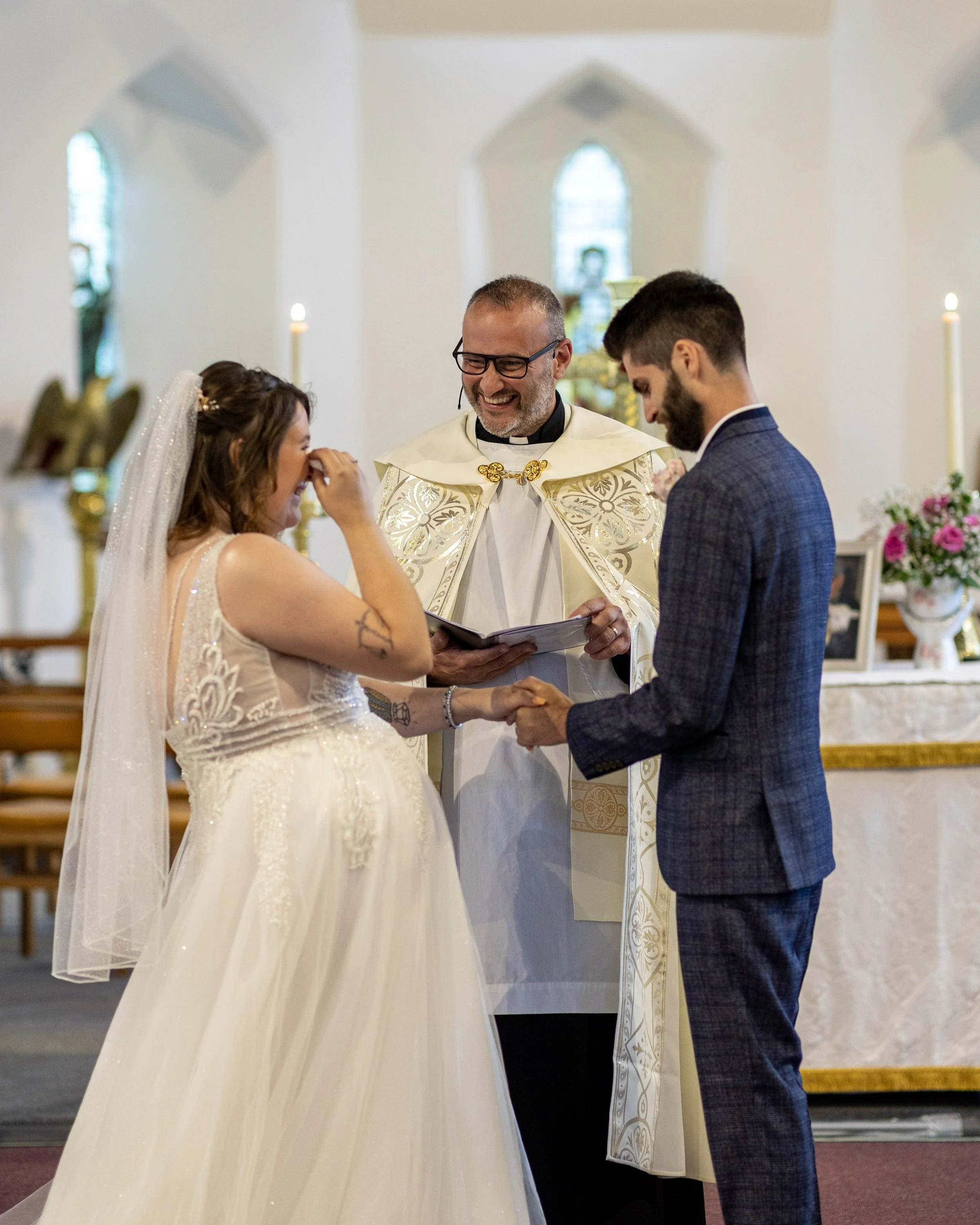 A bride and groom holding hands during their wedding ceremony, with a smiling officiant standing behind them inside a church decorated with flowers and candles.