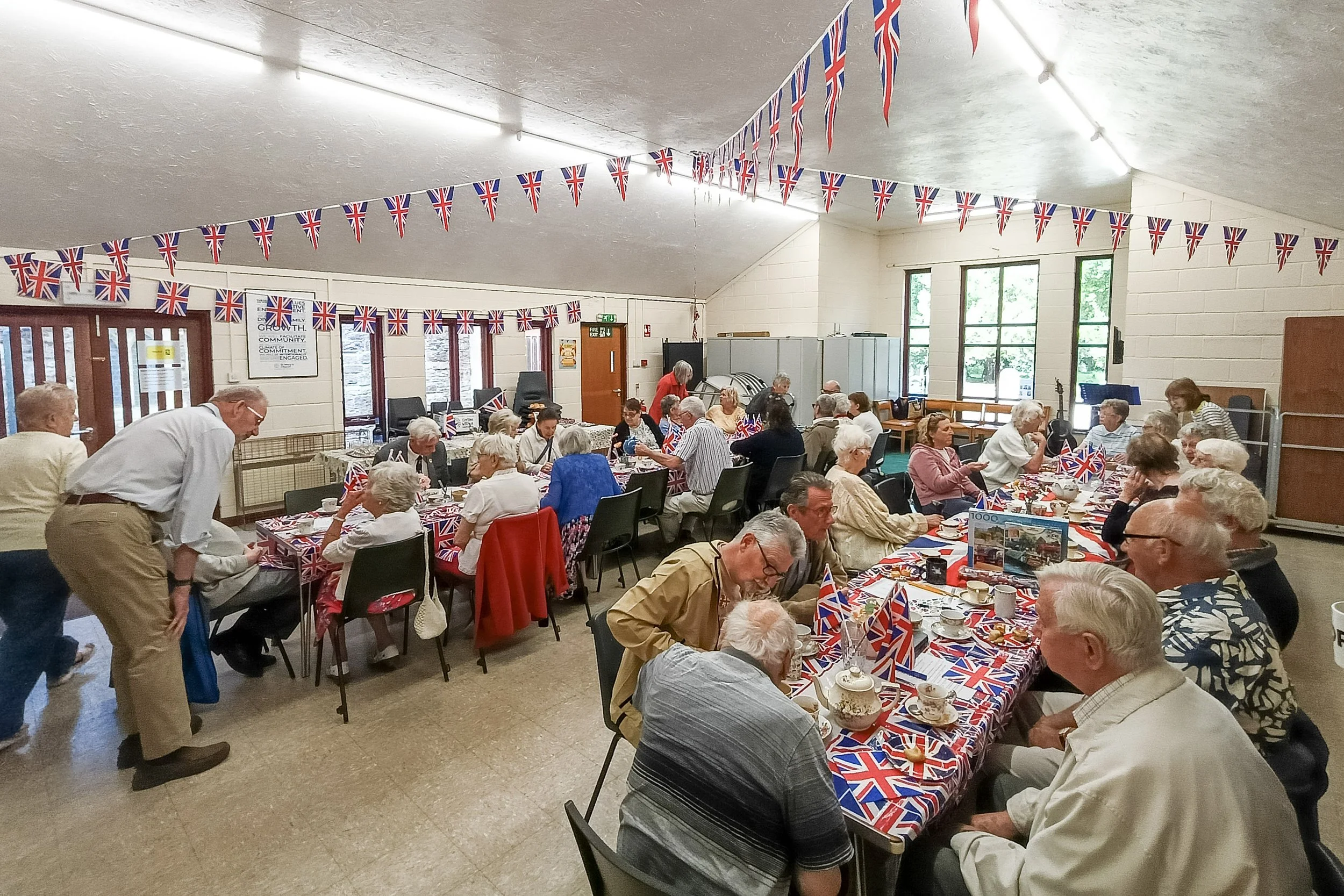 A large gathering of elderly people celebrating with Union Jack flags and decorations, seated around tables covered with patriotic tablecloths, teacups, and teapots, in a community hall decorated for a British-themed event.