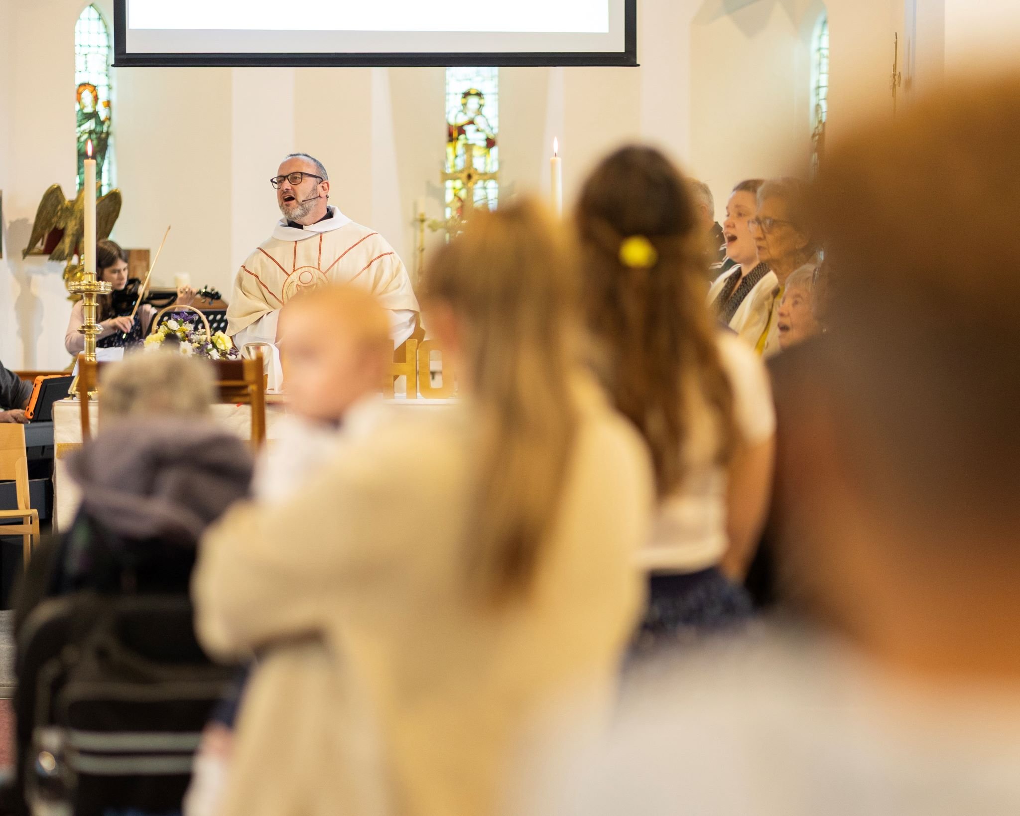A priest leading a church service with choir members singing, several children and adults in attendance, stained glass windows in the background.