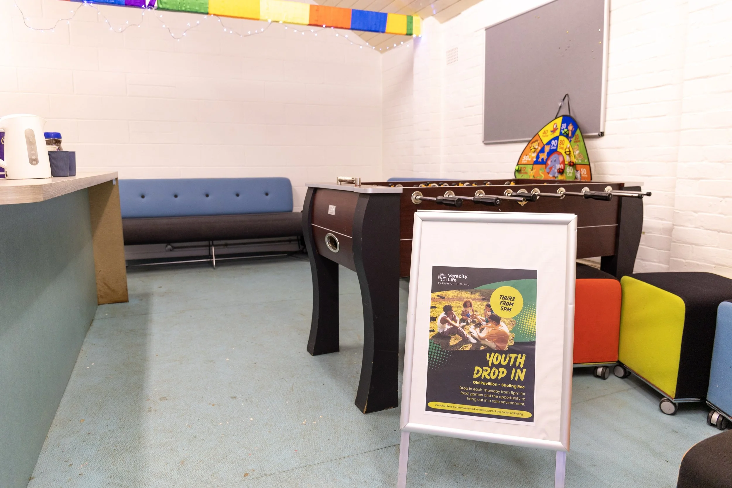 Indoor community space with a foosball table, colorful modular seating blocks, a bench, and a sign promoting youth drop-in activities on Thursday evenings.