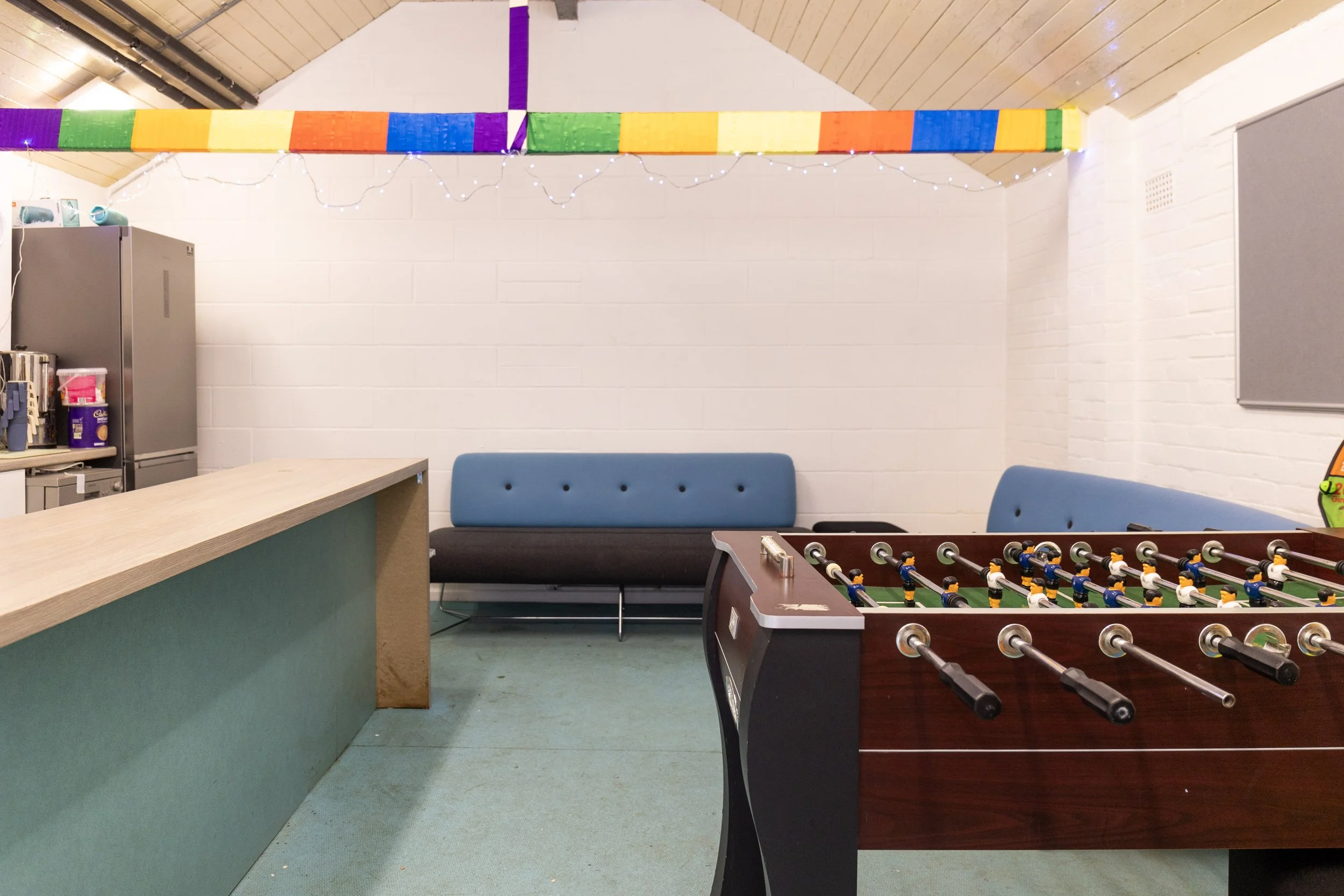 A room decorated with rainbow-colored streamers and string lights, featuring a foosball table, blue cushioned benches, and a counter with snacks and a refrigerator.