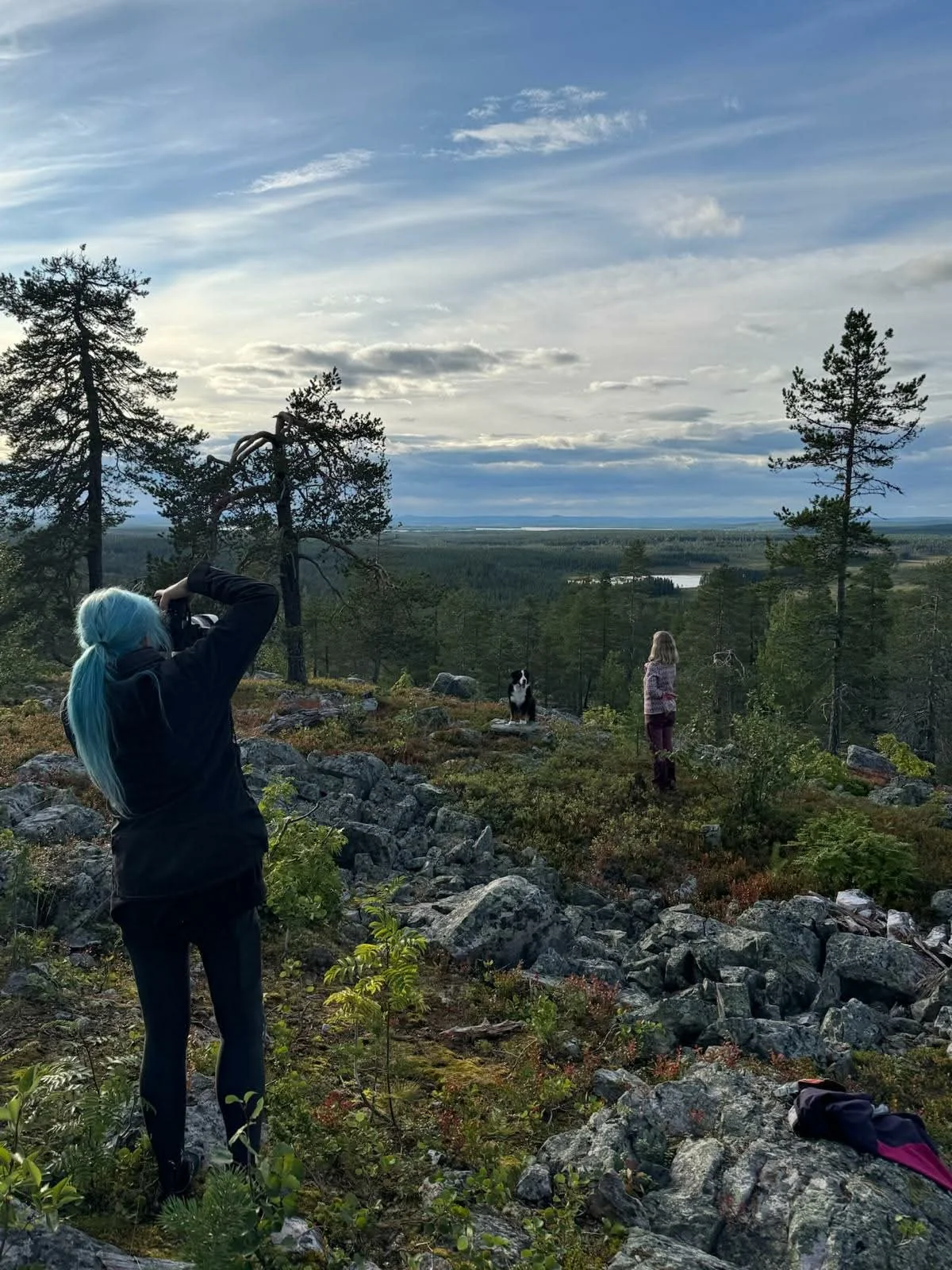 Nainen ottaa valokuvan koirasta, joka istuu kalliolla metsäisessä maisemassa, taustalla järvi ja pilvinen taivas.