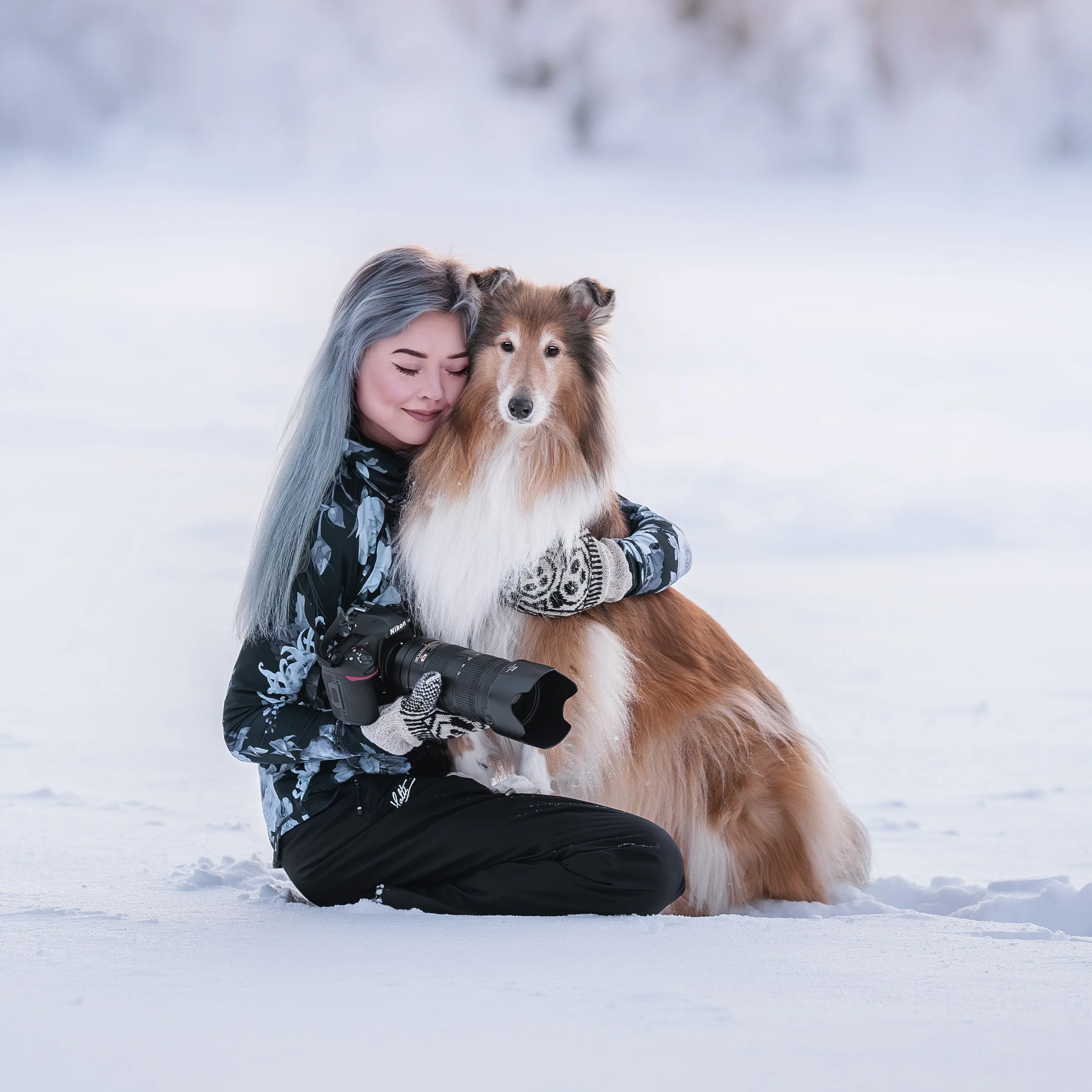 Nainen ja collie-koira talvimaisemassa, nainen pitää kameraa, nainen ja koira halaavat.