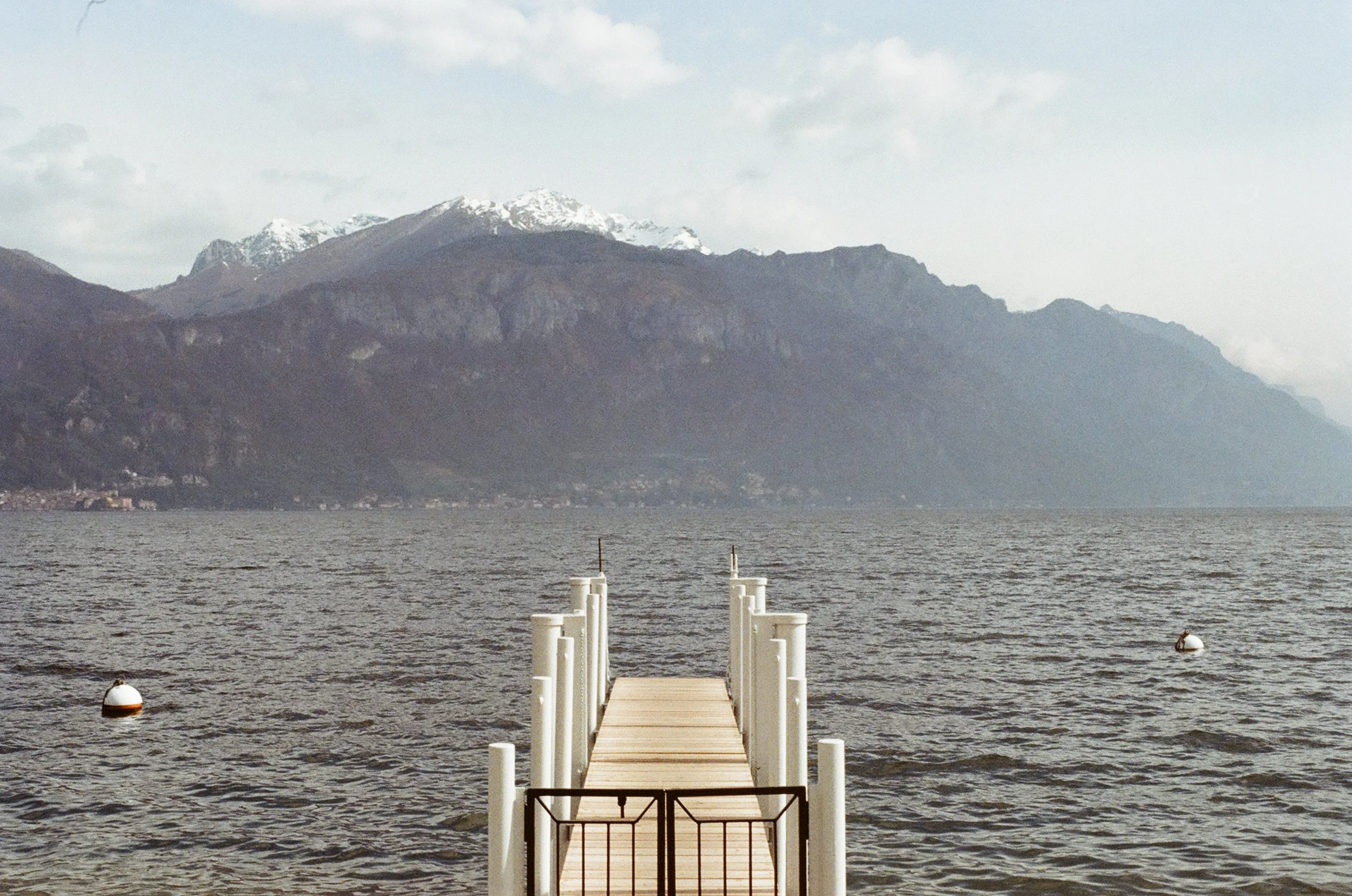 A wooden dock extends into a body of water with mountains in the background, some snow-capped, and a cloudy sky overhead.