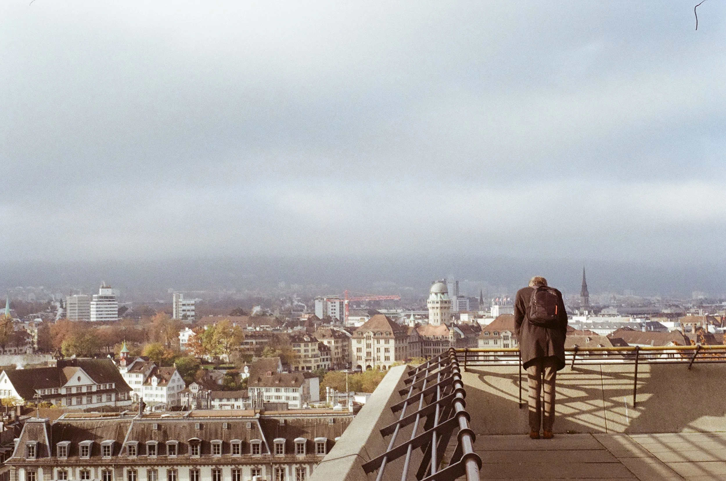 Person with a backpack standing on rooftop observation deck overlooking a cityscape under an overcast sky.