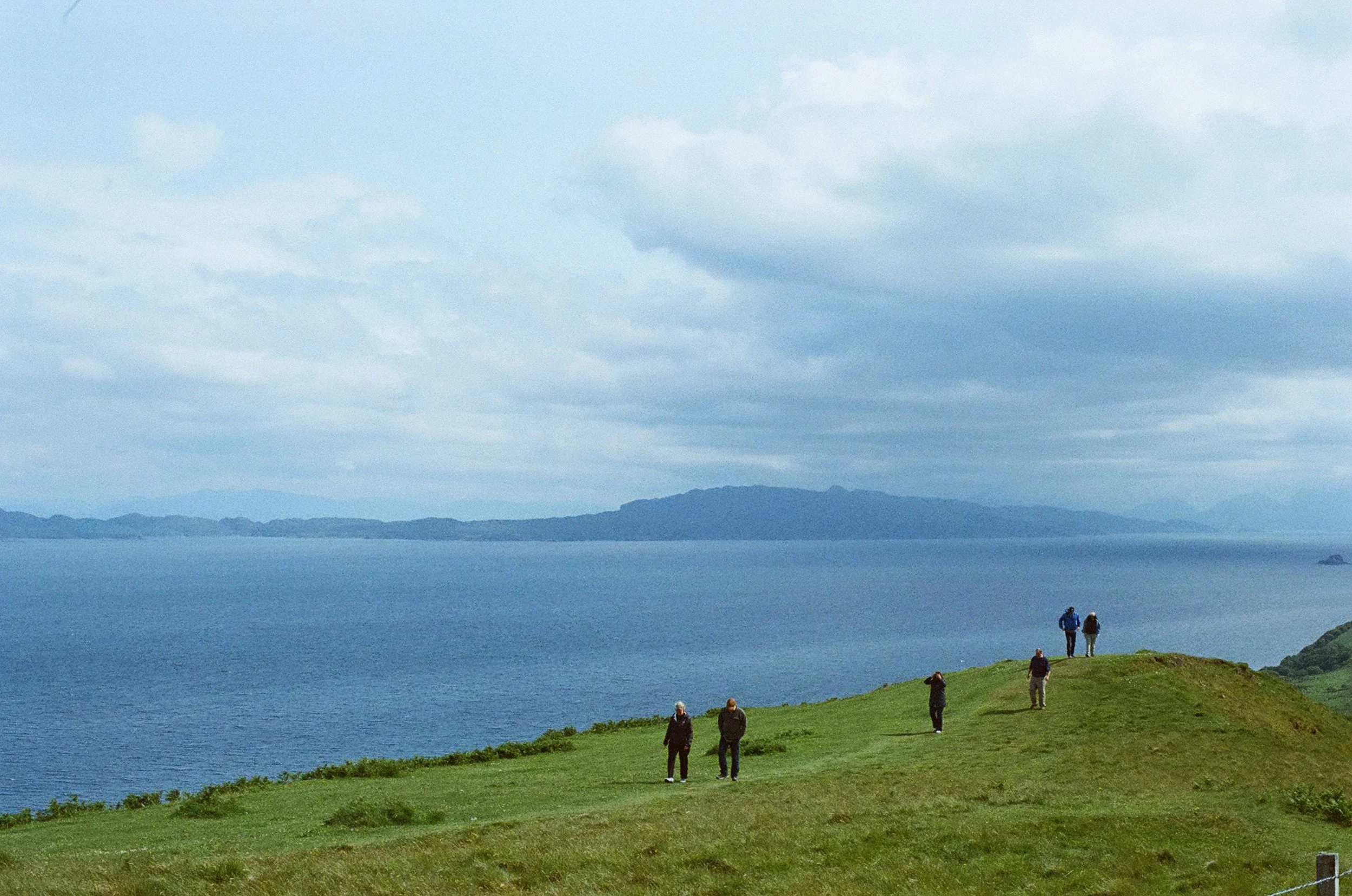 A group of seven people walking and standing on a grassy hill overlooking a body of water and distant islands under cloudy skies.