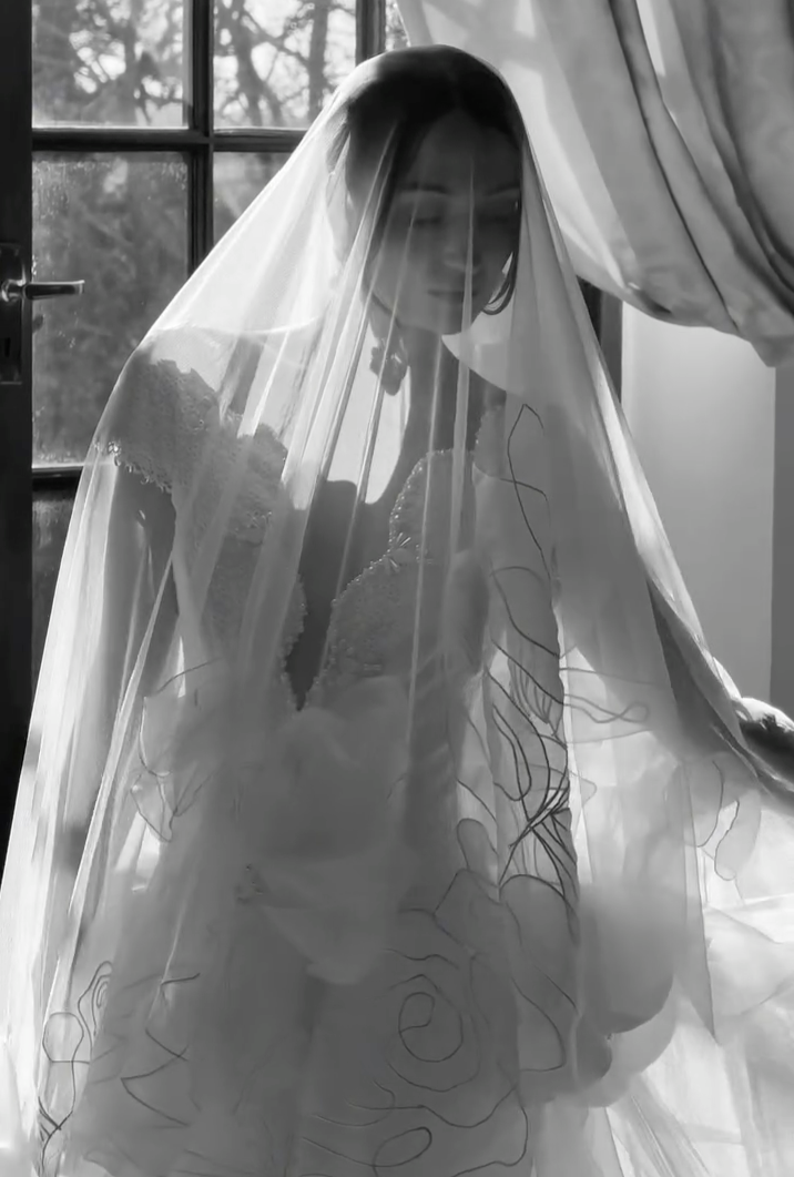 A bride in a wedding dress is seen through a sheer, embroidered veil, sitting by a window with curtains on the side. Natural light filters in, illuminating her face and dress.