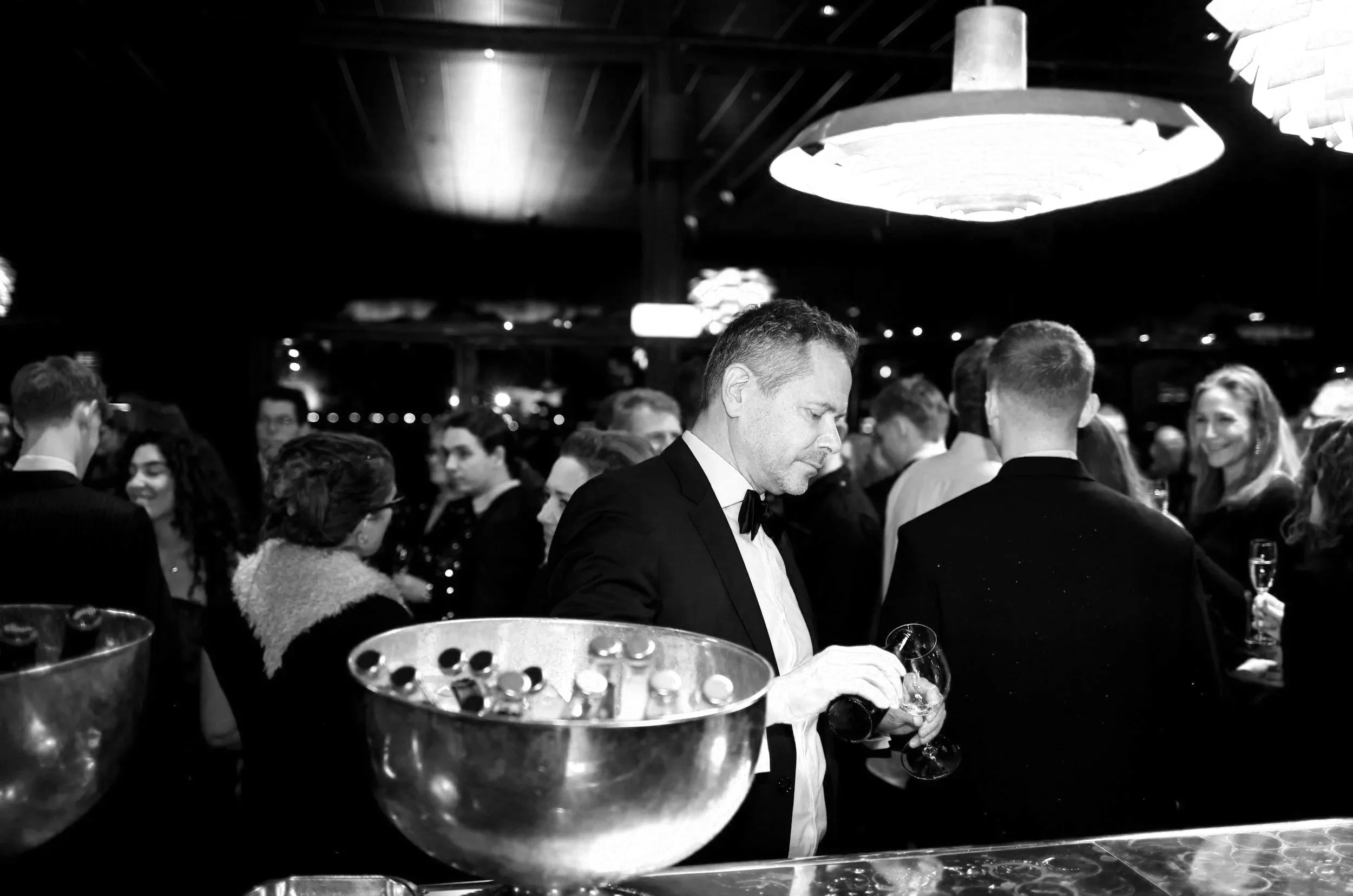 Man in tuxedo with bow tie holding a glass at a party or celebration, surrounded by people in formal attire, with a large light fixture overhead and a reflective surface in the foreground.