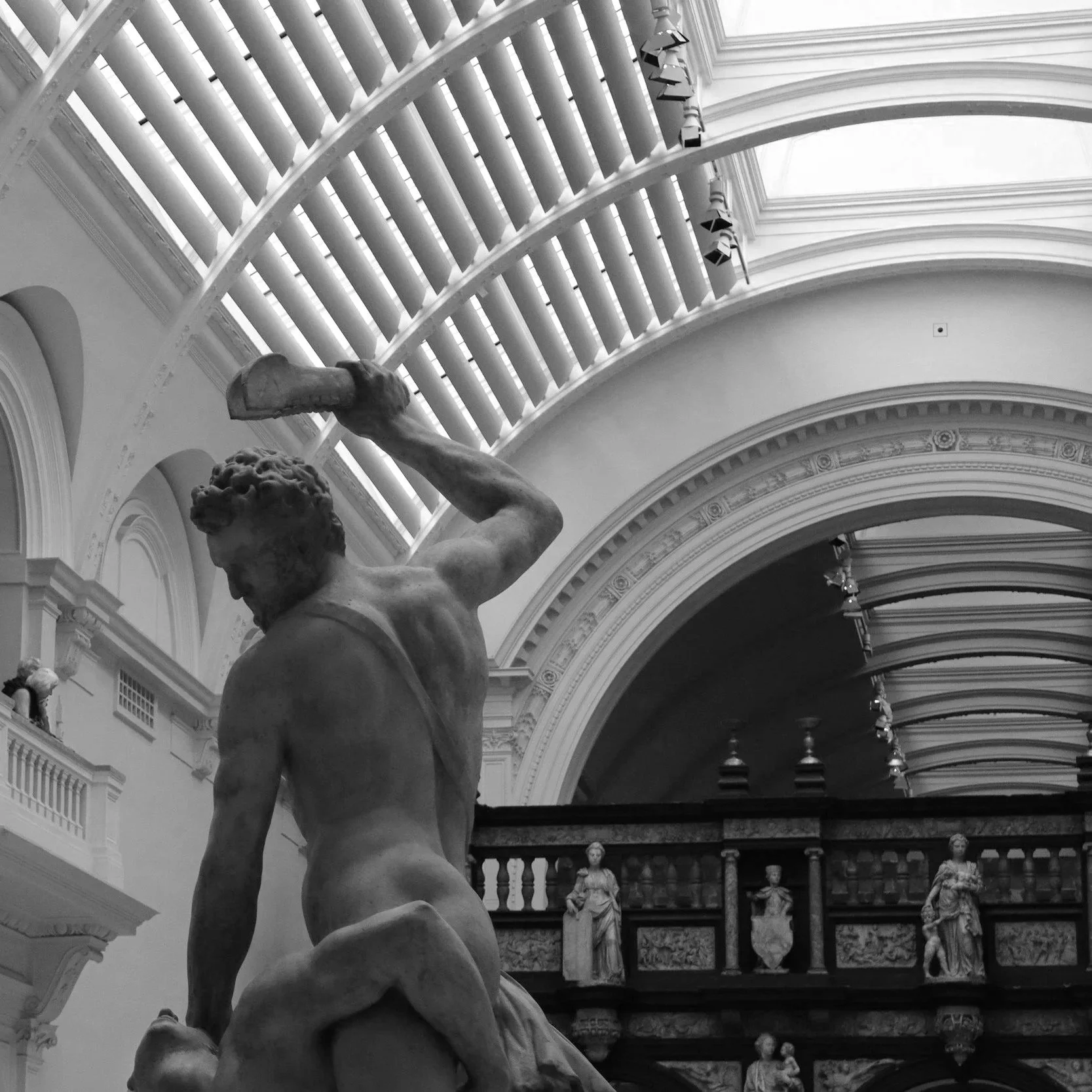 A black and white photograph of a classical marble statue of a muscular male figure holding a hammer, set inside a grand gallery with high, arched ceilings and ornate architectural details.