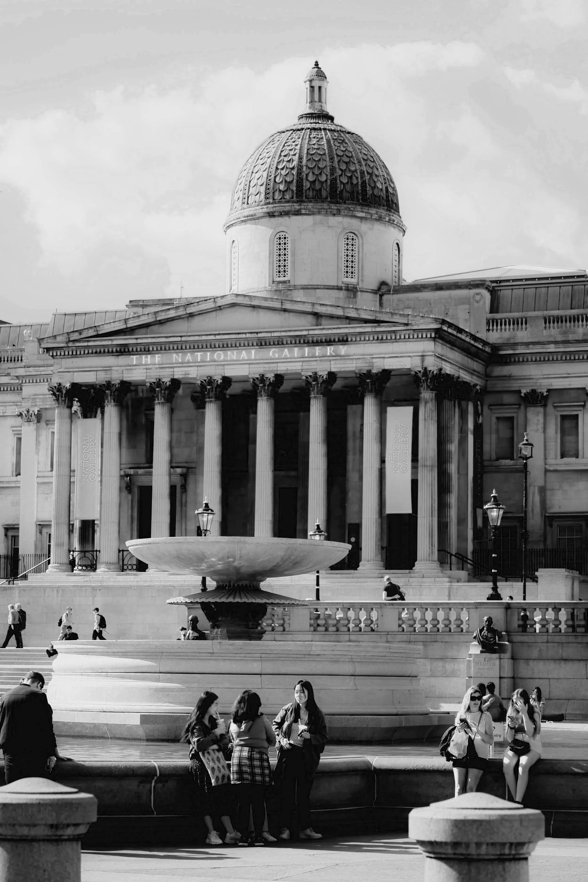 The image shows the front view of the National Gallery in London, located in Trafalgar Square, at the very heart of the city. People sit on the fountains outside in front of the steps up to the columns lining the entrance