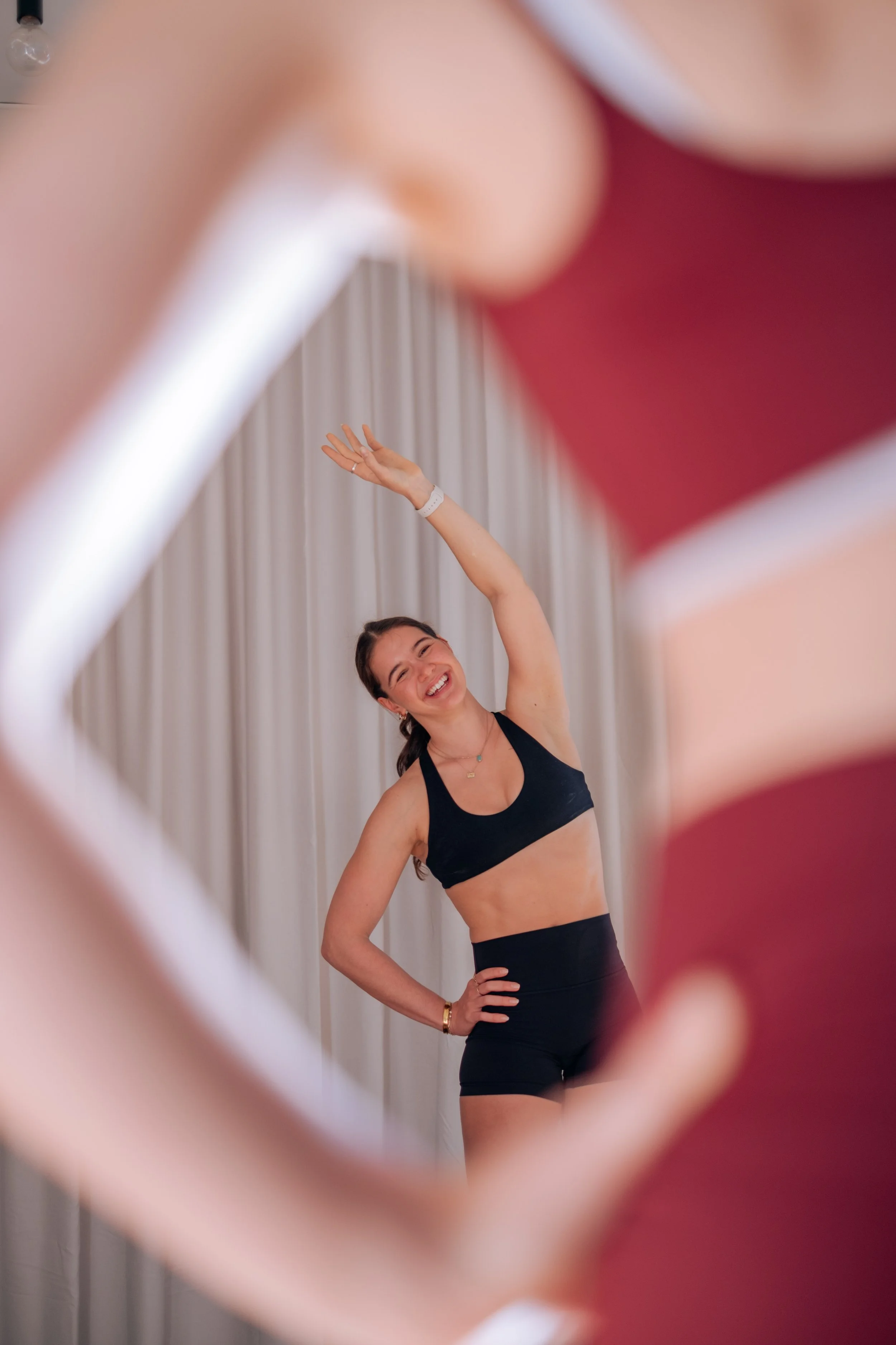 A woman in a black sports bra and black shorts is smiling and posing with her hand on her hip, seen through a pink heart-shaped object in the foreground.