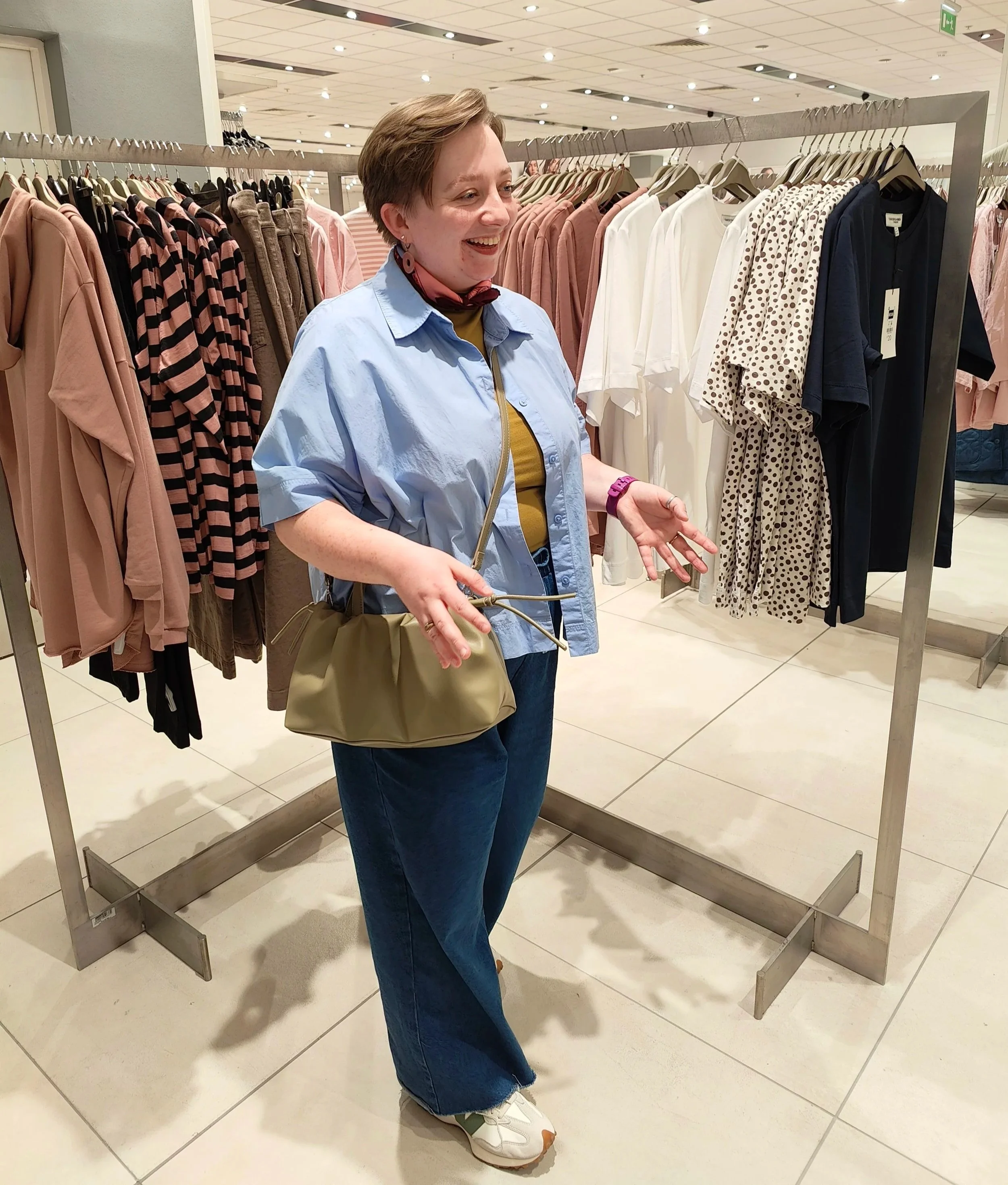 A stylist laughing while standing between clothing rails, wearing a blue button-down shirt, olive top, and wide-leg jeans, with a small olive crossbody bag.