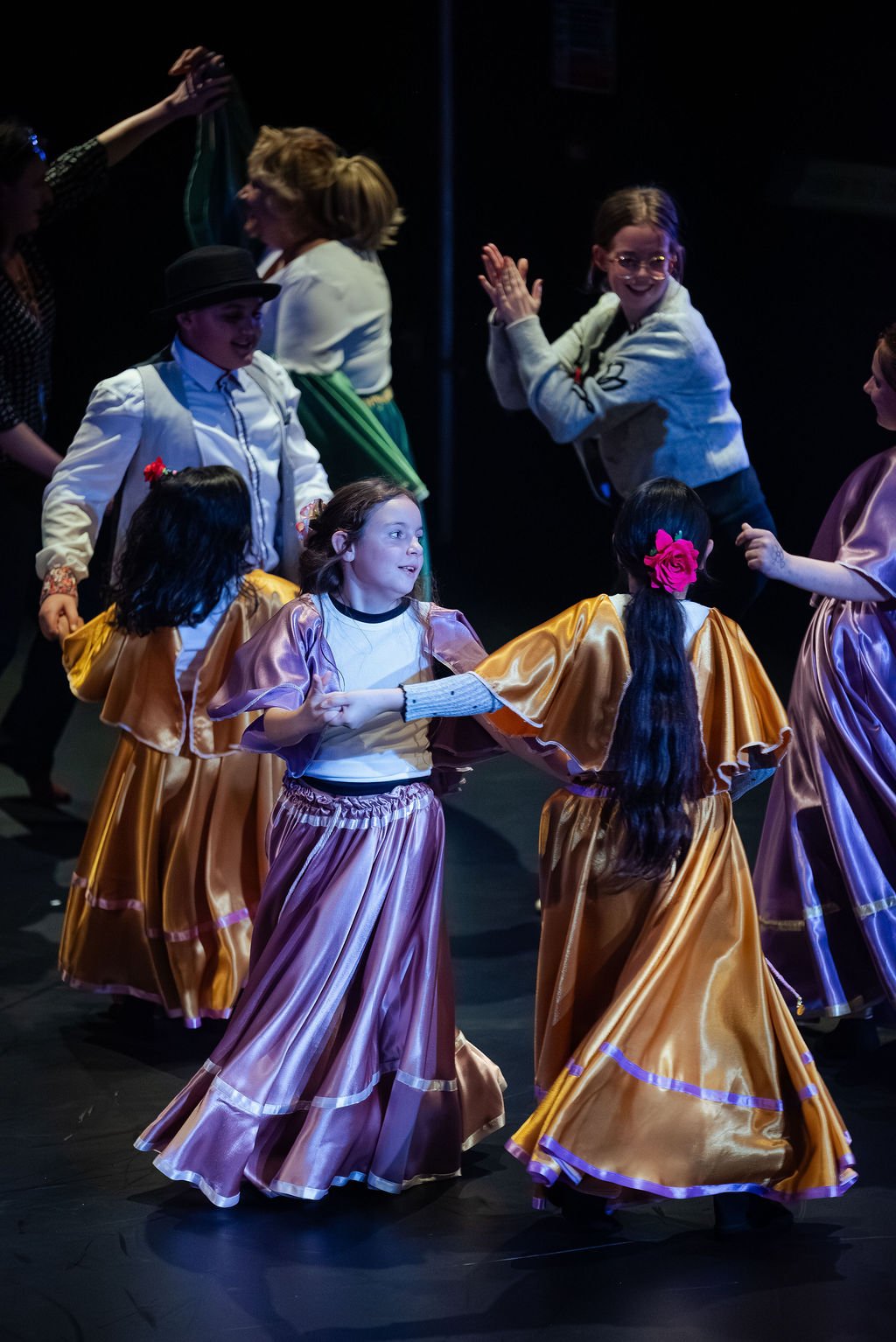 Children in gold and purple satin skirts dance in a circle on stage, adults performing behind them, in MOV Dance Theatre's Voices performance.