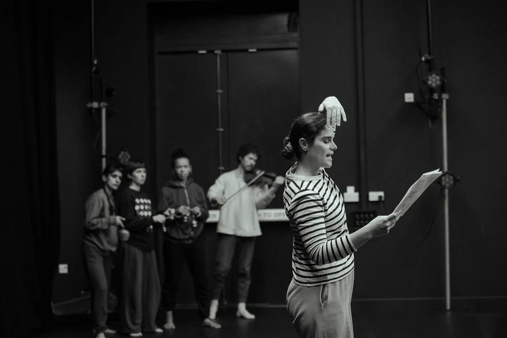 OV Dance Theatre performer holds script with hand raised to forehead, ensemble and violinist visible behind in studio rehearsal.