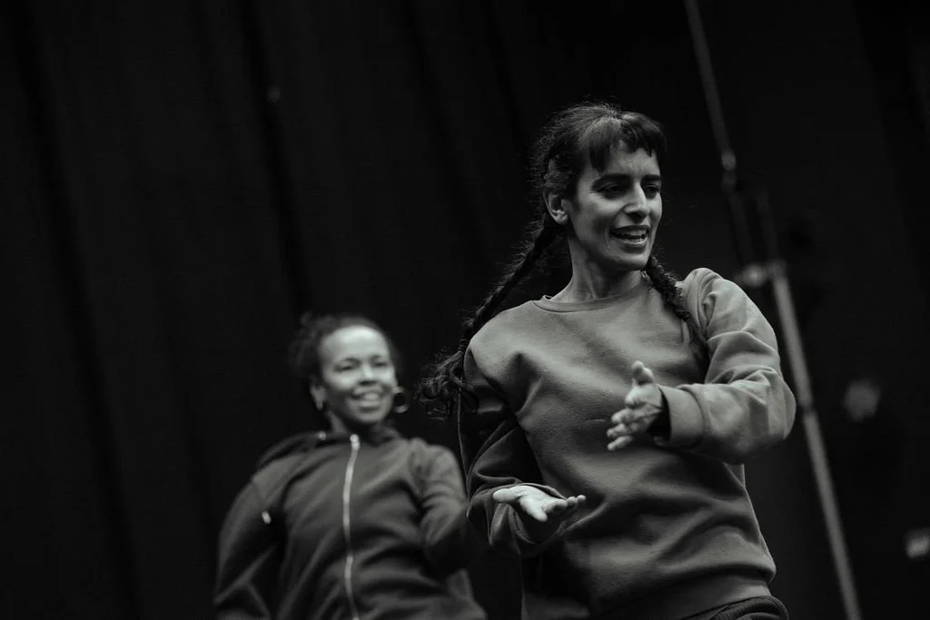 Two MOV Dance Theatre performers mid-movement in black and white rehearsal shot, smiling, against dark stage drapes.