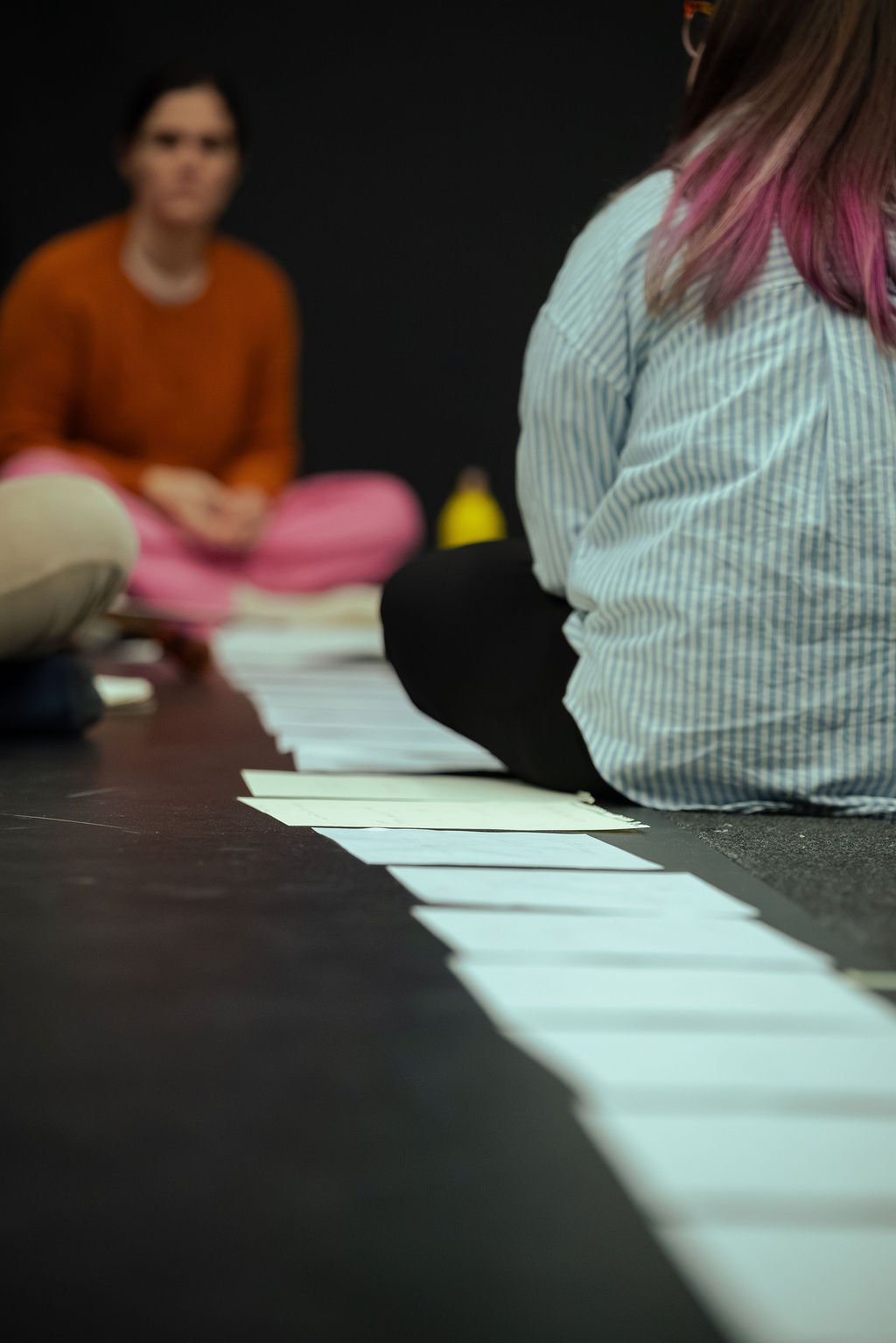 Two performers sit on a dark studio floor either side of a line of white cards laid between them in a MOV Dance Theatre rehearsal process shot.