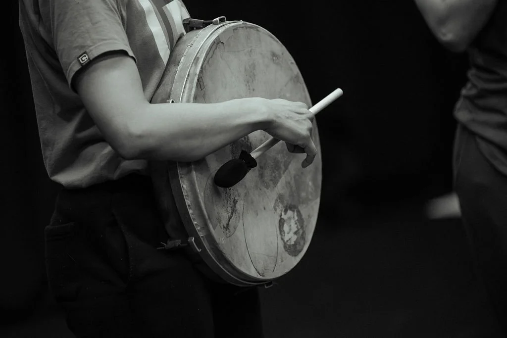 Close crop of MOV Dance Theatre performer holding and playing a frame drum with a beater during rehearsal.