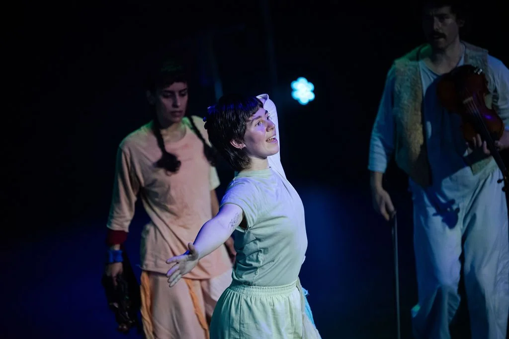 Dancer in a white dress extends her arms on a blue-lit stage, a violinist and another performer behind her in a MOV Dance Theatre performance.