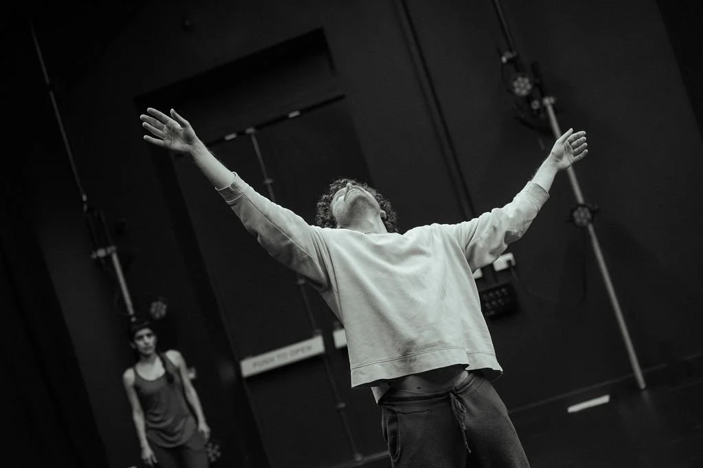 MOV Dance Theatre male performer stretches arms wide overhead, face tilted up, second dancer watching from background in studio.