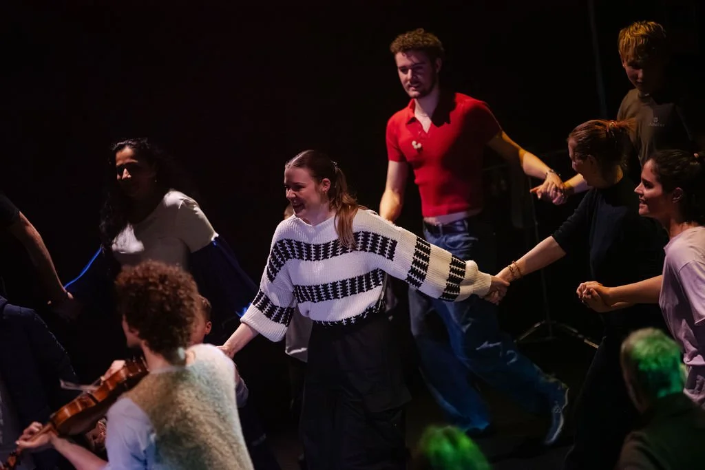 Ensemble cast of MOV Dance Theatre's Voices hold hands on a darkly lit stage, smiling, with a violinist visible in the foreground.