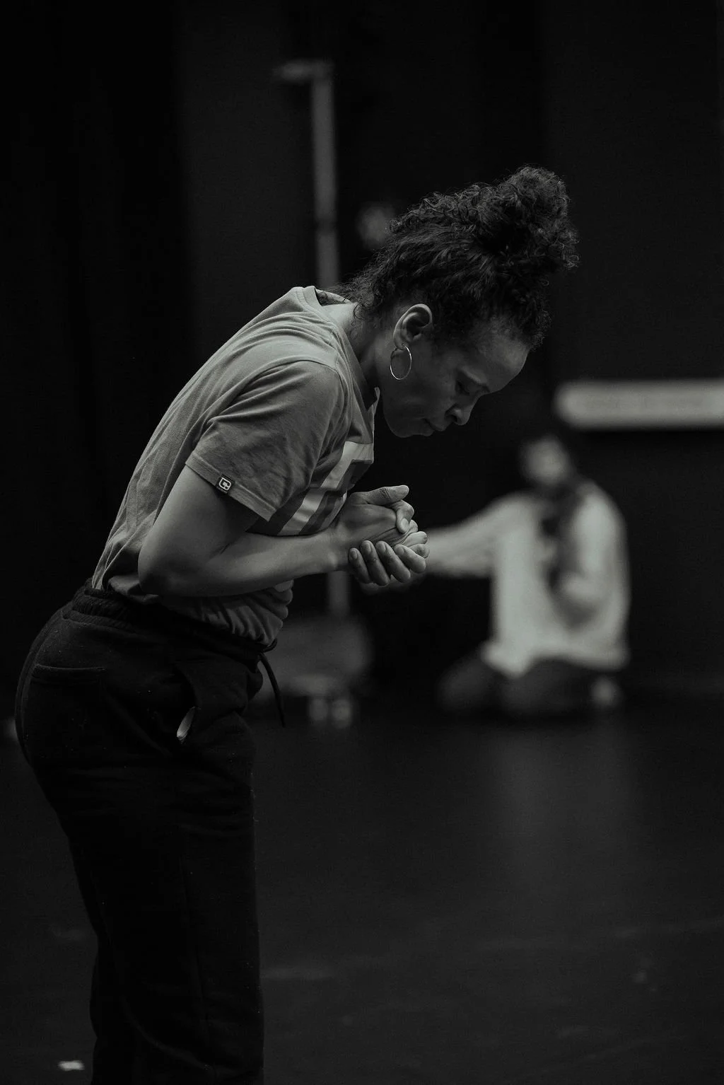 MOV Dance Theatre dancer bends forward with clasped hands in focused rehearsal, second performer seated in soft focus behind.