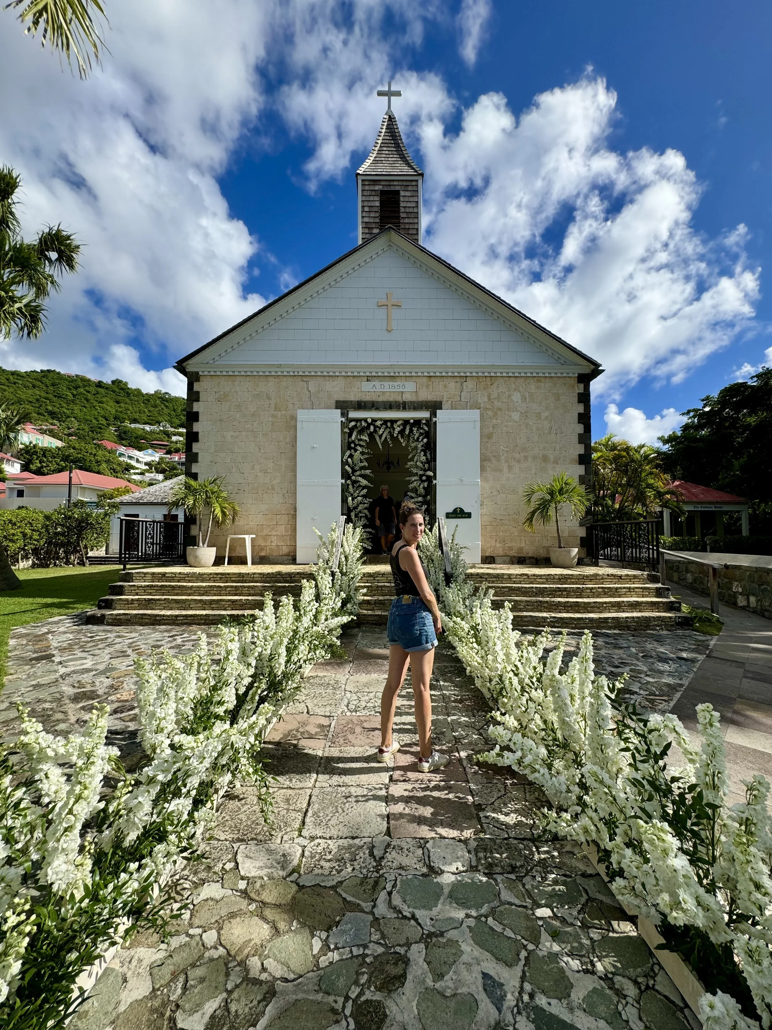 Une jeune femme se tient devant une petite église blanche en pierre, entourée de fleurs blanches, avec un ciel bleu et quelques nuages en arrière-plan.