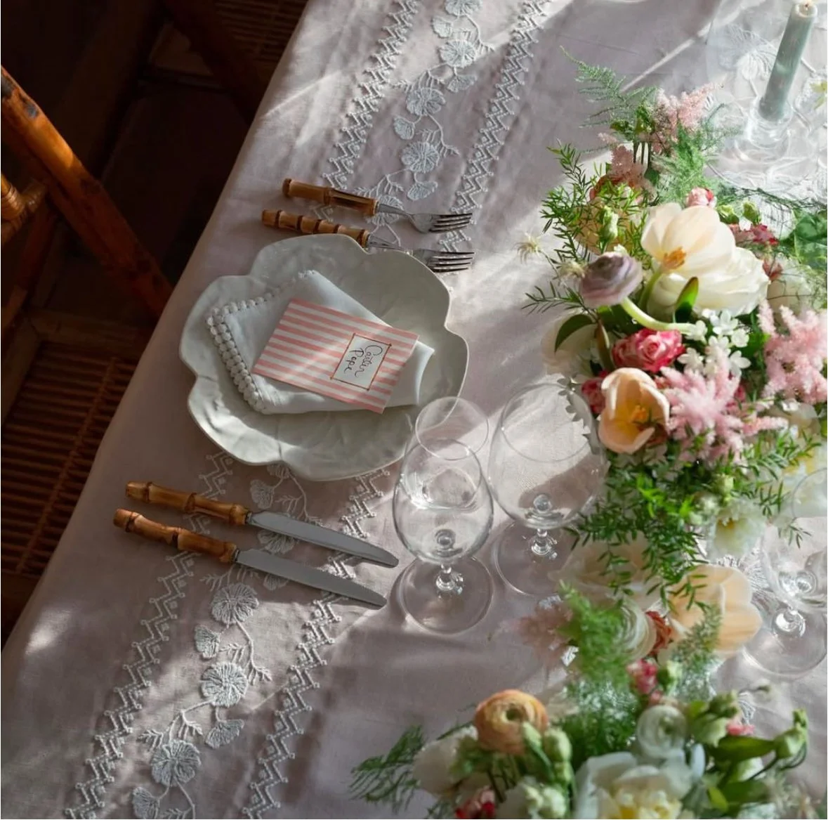 Table dressée pour un repas avec une nappe blanche, des serviettes pliées, des fourchettes et couteaux à manches en bois, des verres à vin, une grande composition florale avec des fleurs blanches, roses et vertes et une bougie.