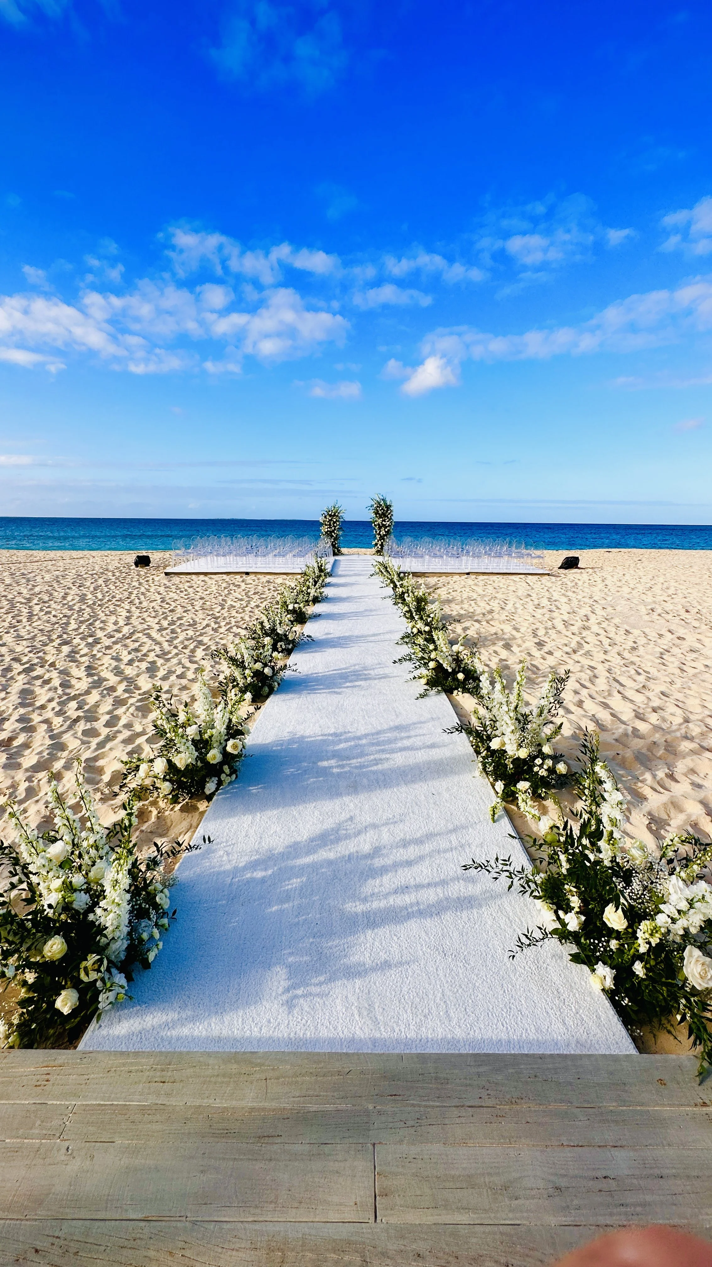 Allée de mariage avec des fleurs blanches sur une plage, face à la mer, sous un ciel bleu avec quelques nuages.