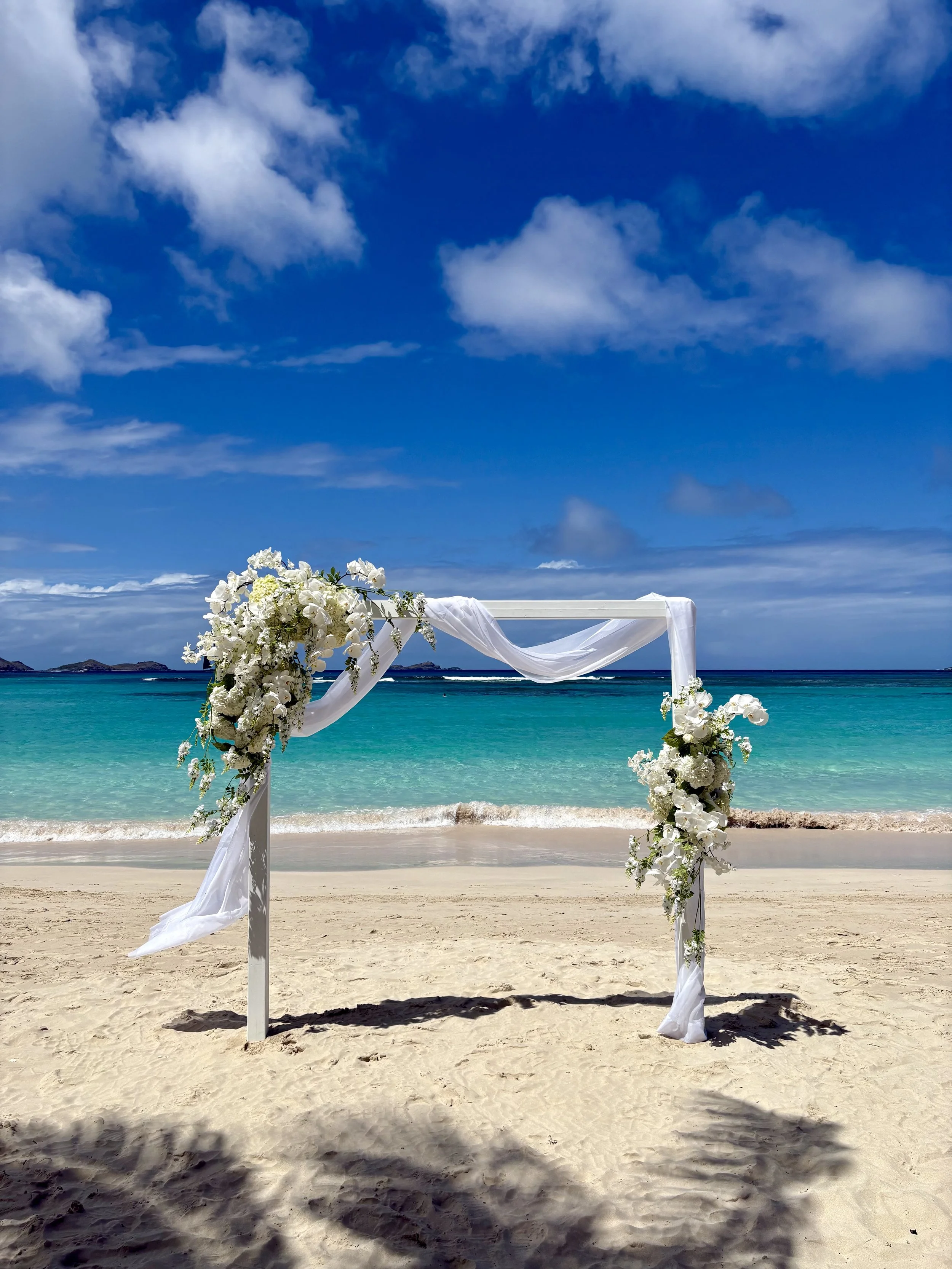 Arche avec décoration florale sur la plage, vue sur mer turquoise et ciel bleu avec nuages