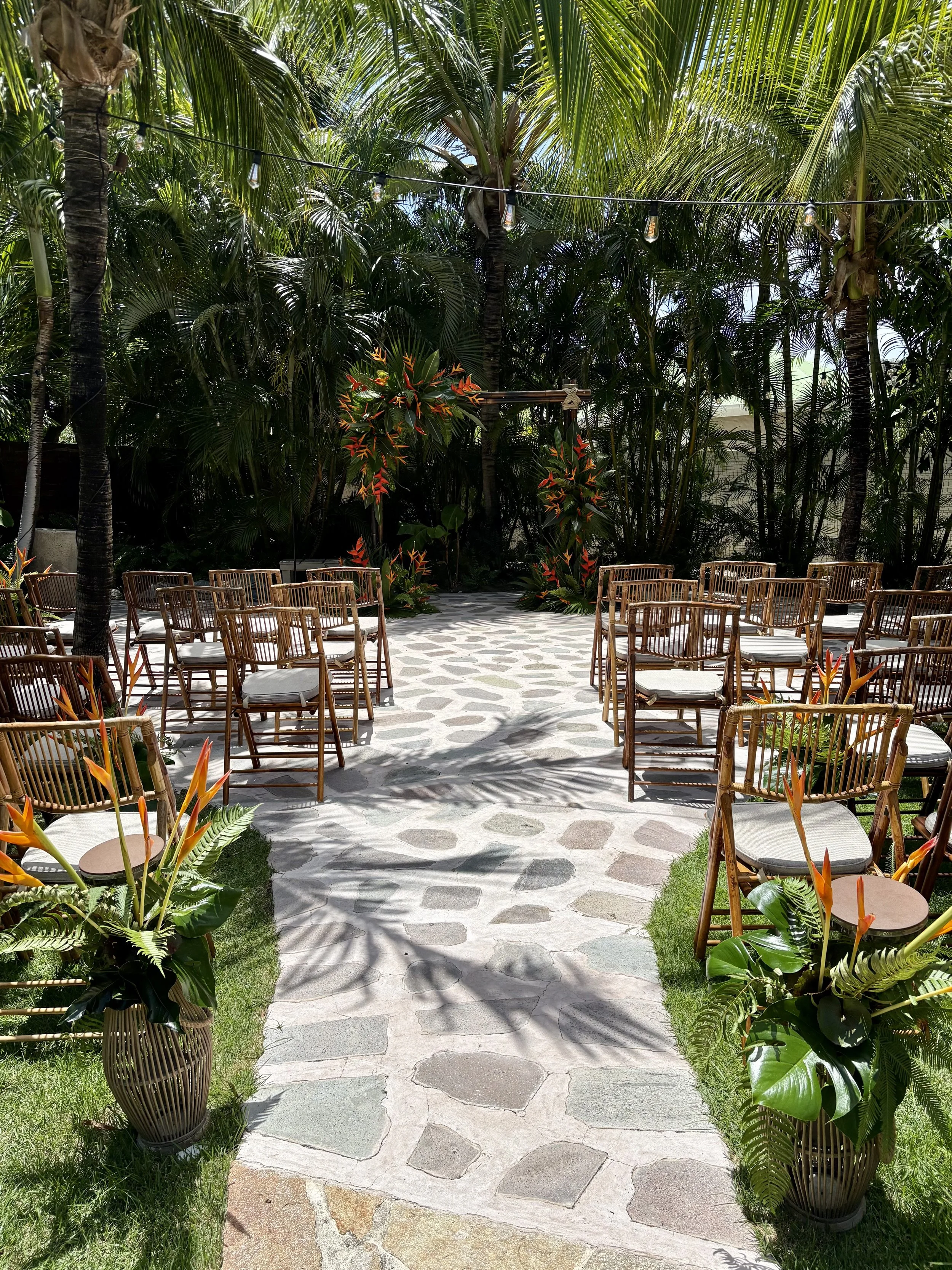 Une salle en plein air pour une cérémonie avec des chaises en bois, des fleurs tropicales orange et vertes, et une toile de fond de palmiers luxuriants.