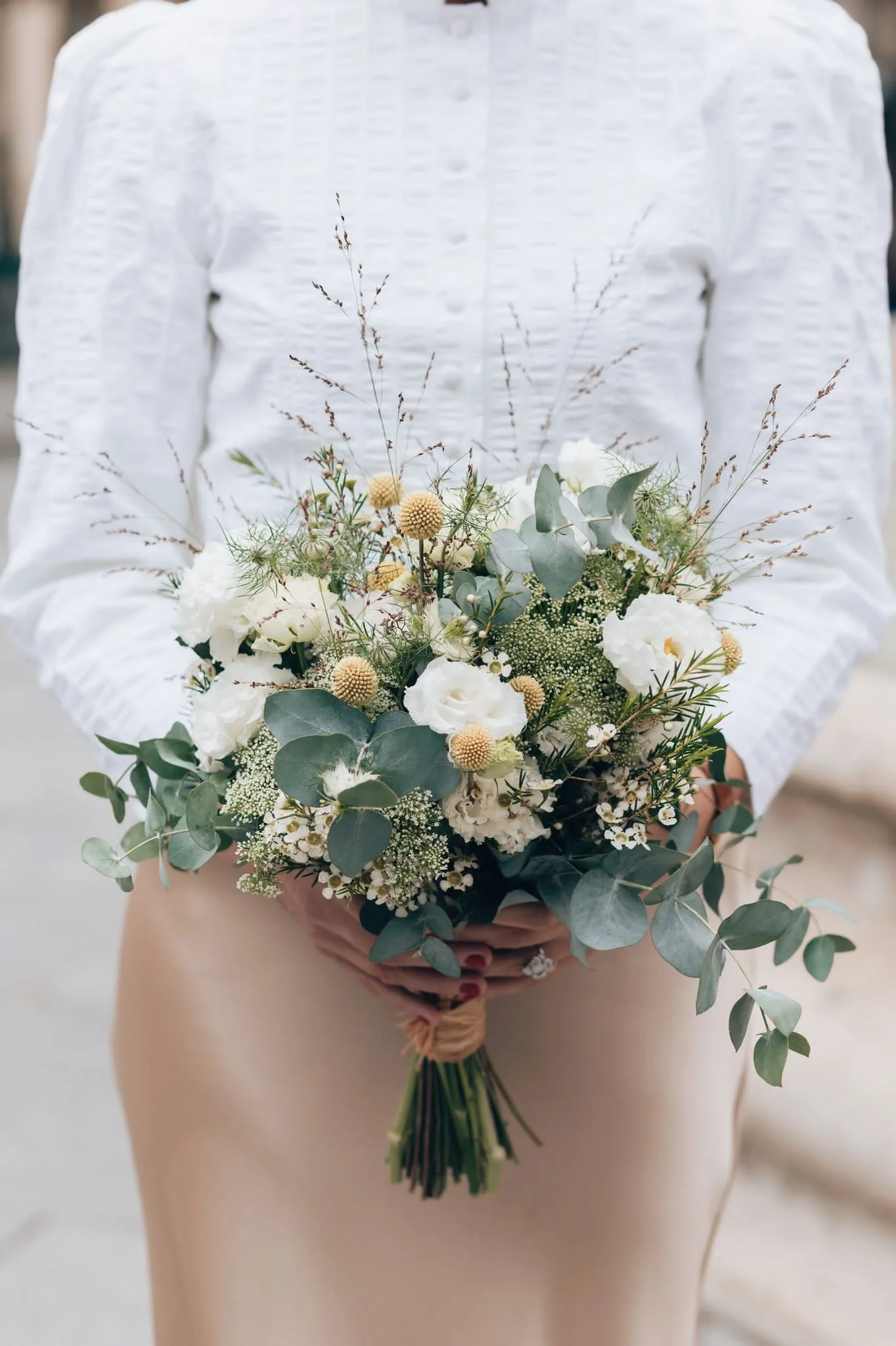 Une mariée tient un bouquet de fleurs blanches et vertes avec des feuillages eucalyptus et petites fleurs, habillée d'une chemise blanche et d'une jupe beige.
