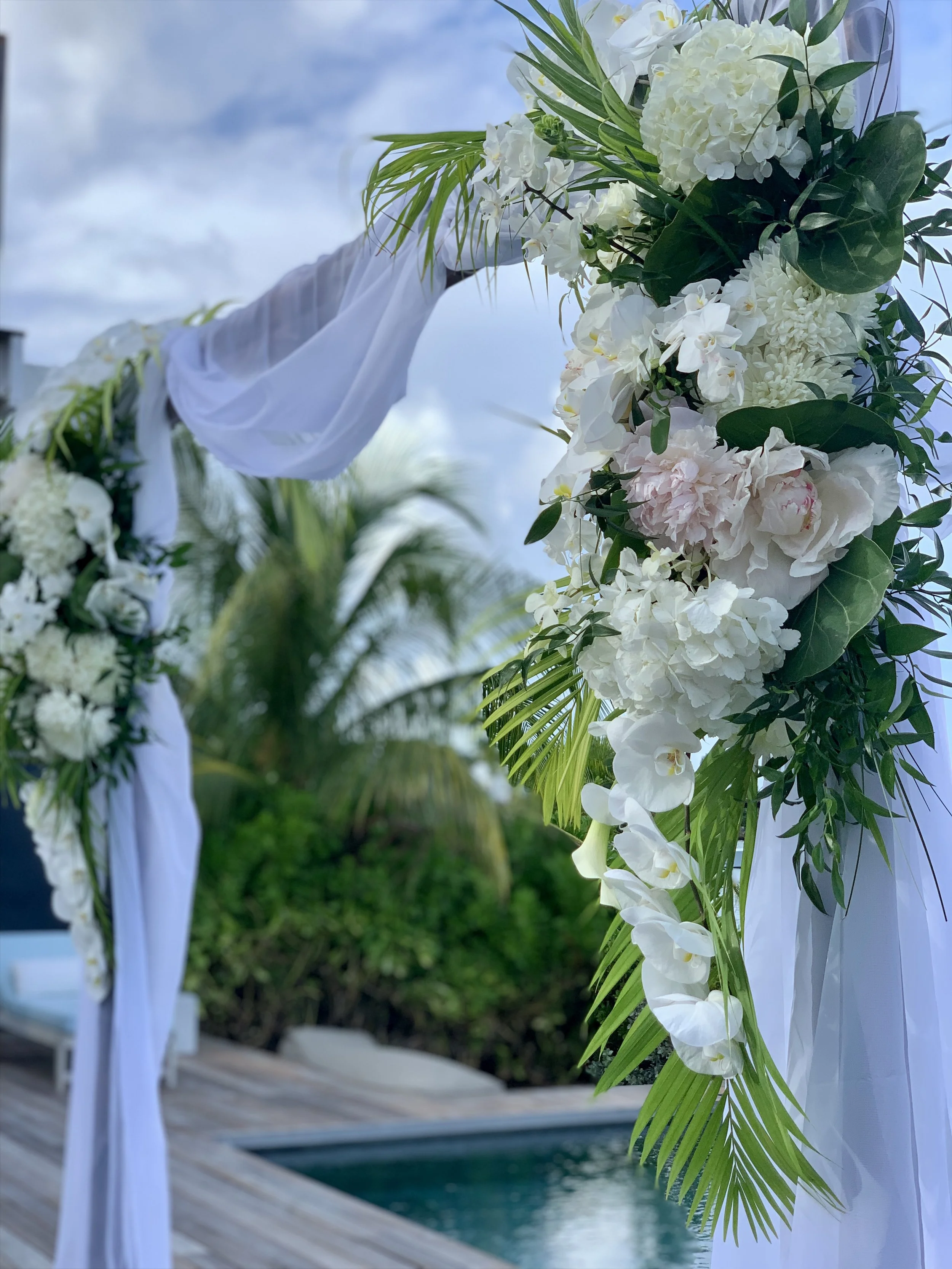 Décor de mariage avec une arche en fleurs blanches et feuilles vertes près d'une piscine, avec un ciel nuageux en arrière-plan.