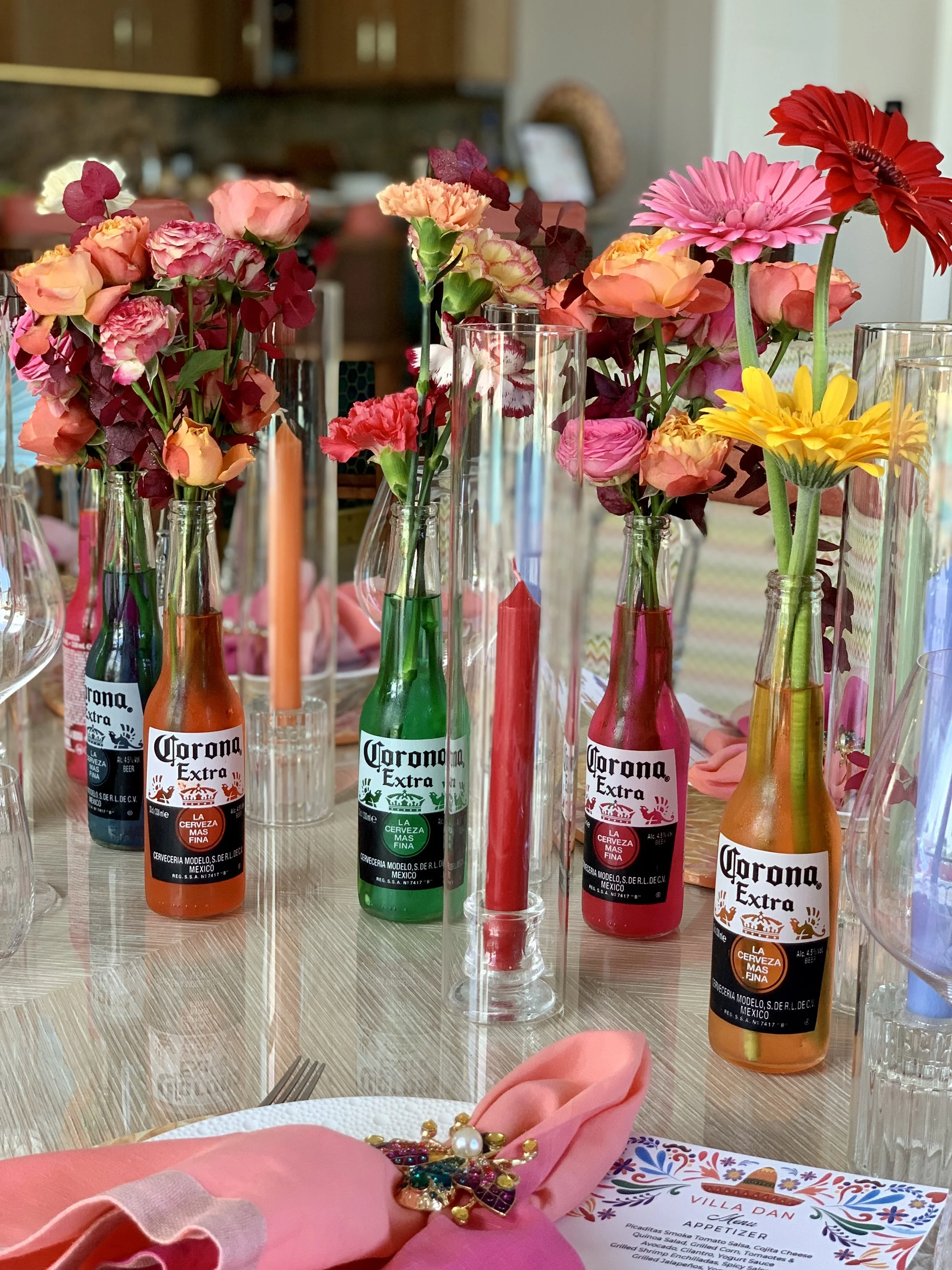 Table decorée avec des bouteilles de Corona en tant que vases, contenant des fleurs colorées, et un centre de table avec des bougies. Présence de nappes roses, un menu, et des décorations de table.