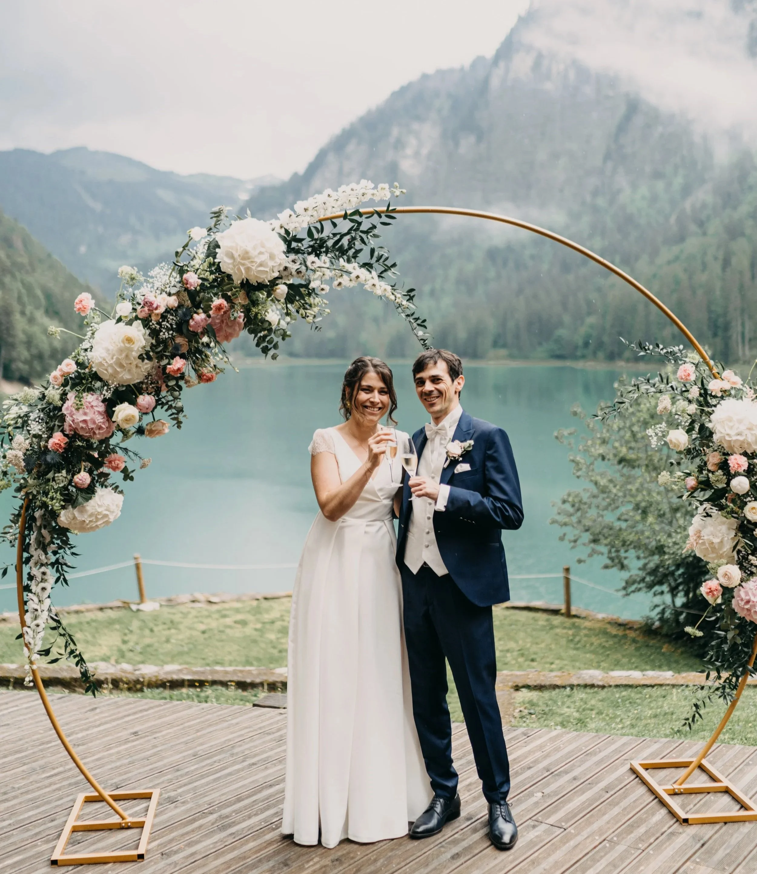 Un couple de mariés souriants, portant des tenues de mariage, se tient sous une arche florale au bord d'un lac entouré de montagnes et de forêts.