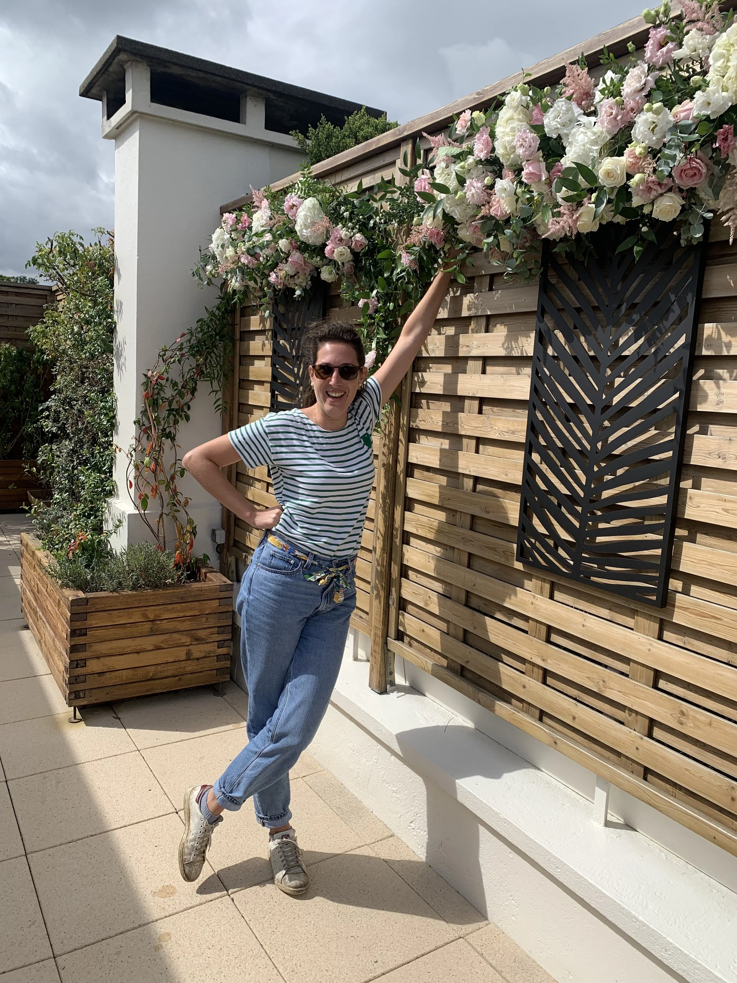 Femme souriante portant un t-shirt rayé et un pantalon en jeans, levant un bras vers des fleurs roses et blanches sur un mur en bois