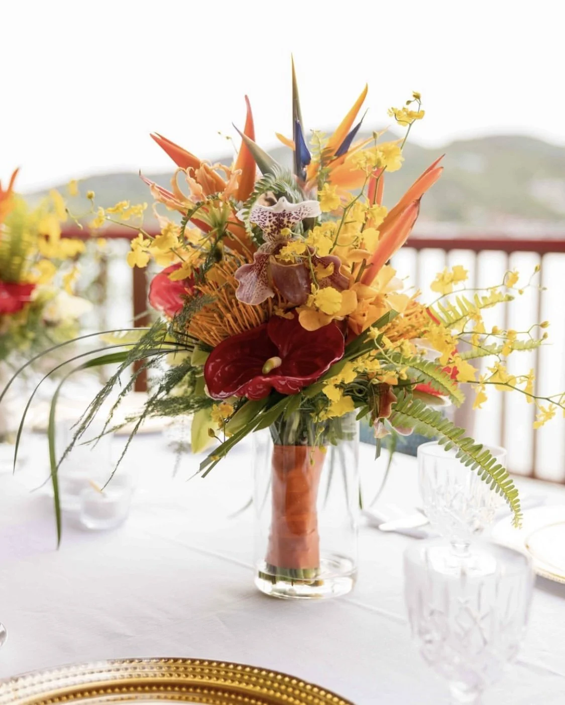 Un bouquet de fleurs tropicales colorées dans un vase en verre sur une table, avec des éléments de décoration et des verres en cristal.
